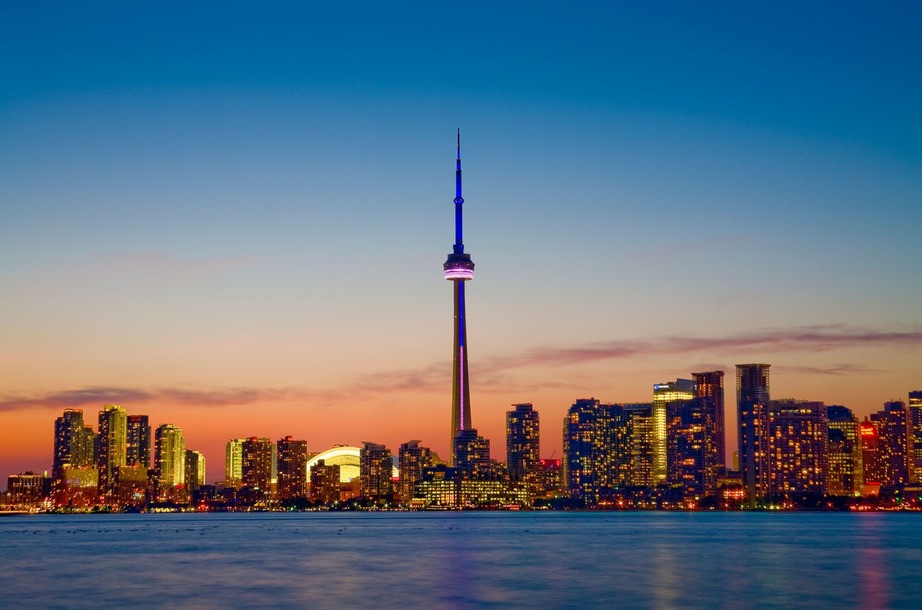 A cityscape lighting up at dusk along a smooth, blue, body of water