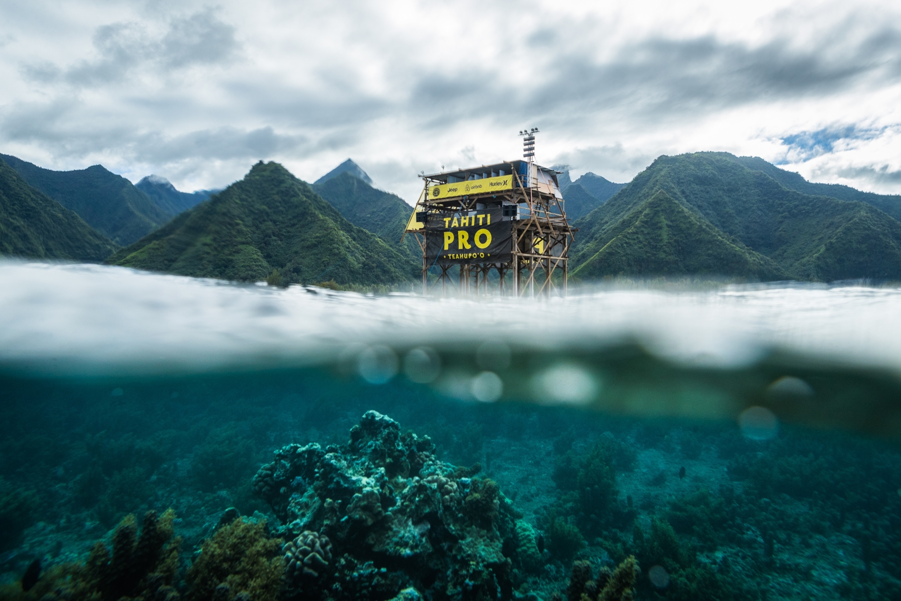 A partially submerged view of a multi-level, wooden judging tower, visible above the water, while rocky coral is visible beneath the water.