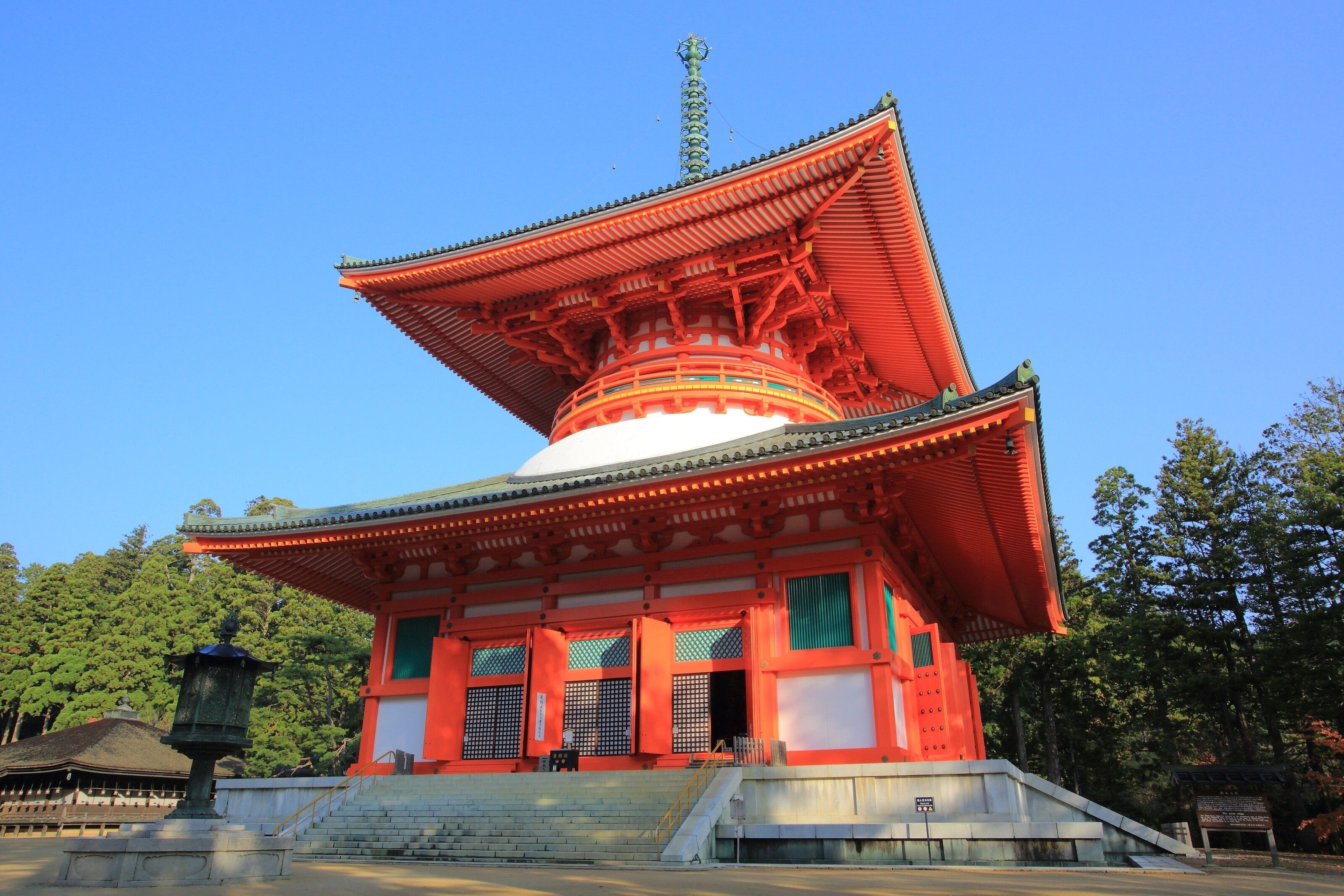 A fiery red temple. It is square, and has a roof for each of its two stories. At the top, a turquoise mast. Forest trees are in the background, and the sky is blue.