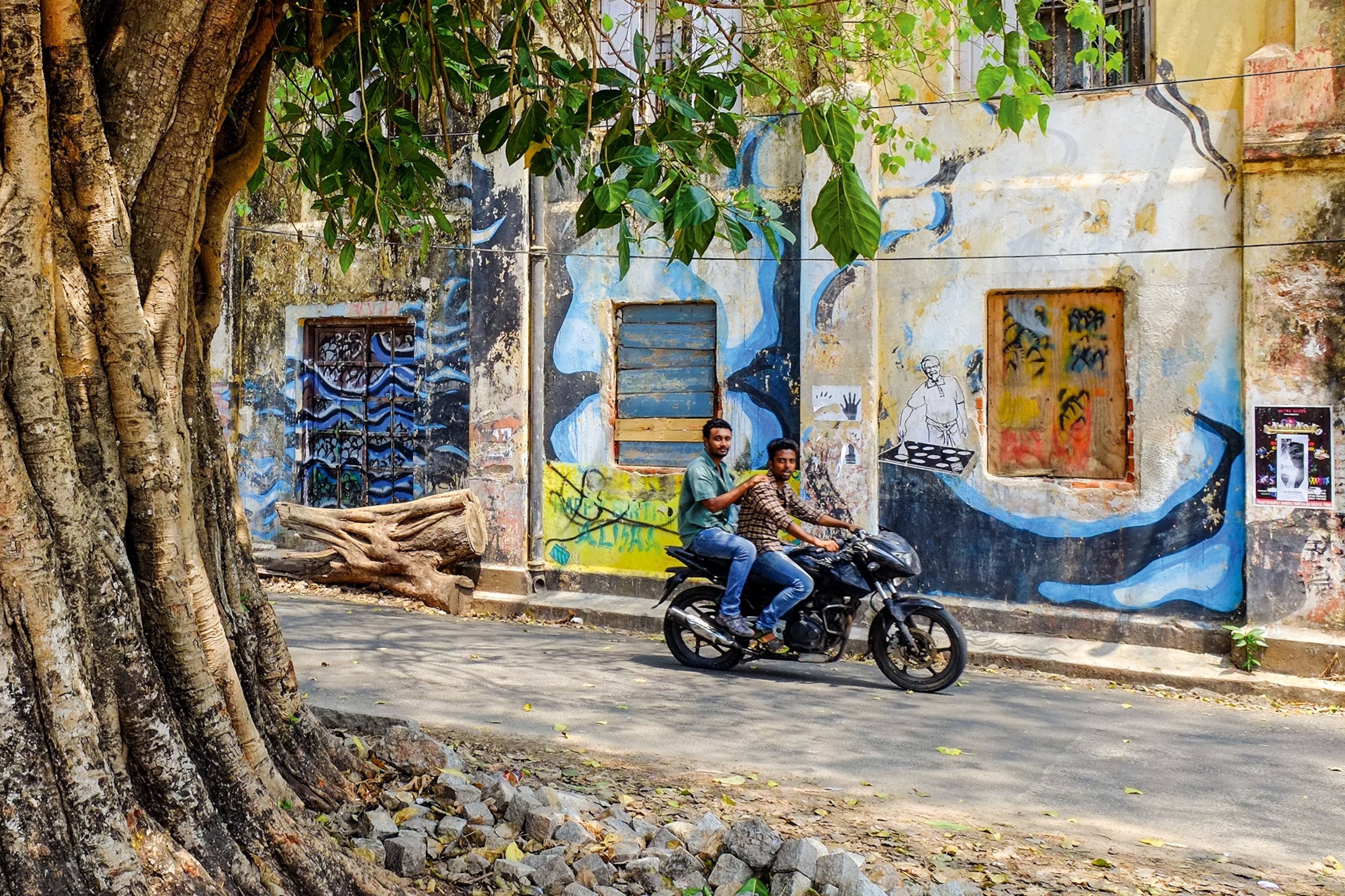 Two Indian men driving by an ancient tree on a motorbike, illustrated house walls behind them.