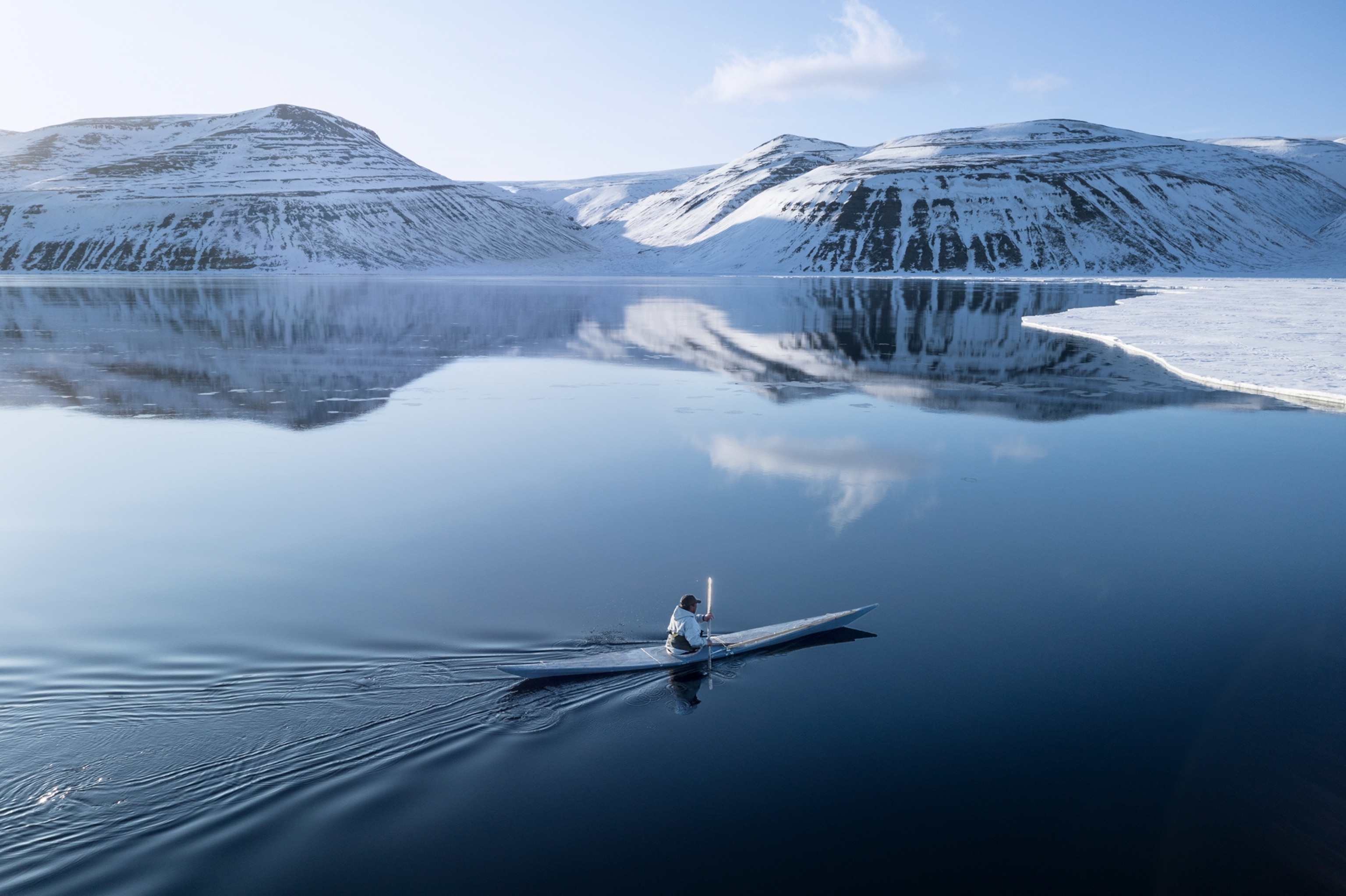 Mikile Kristiansen is kayaking toward the icy shore on calm water. Mountains are behind him, and there are a few clouds in the sky.