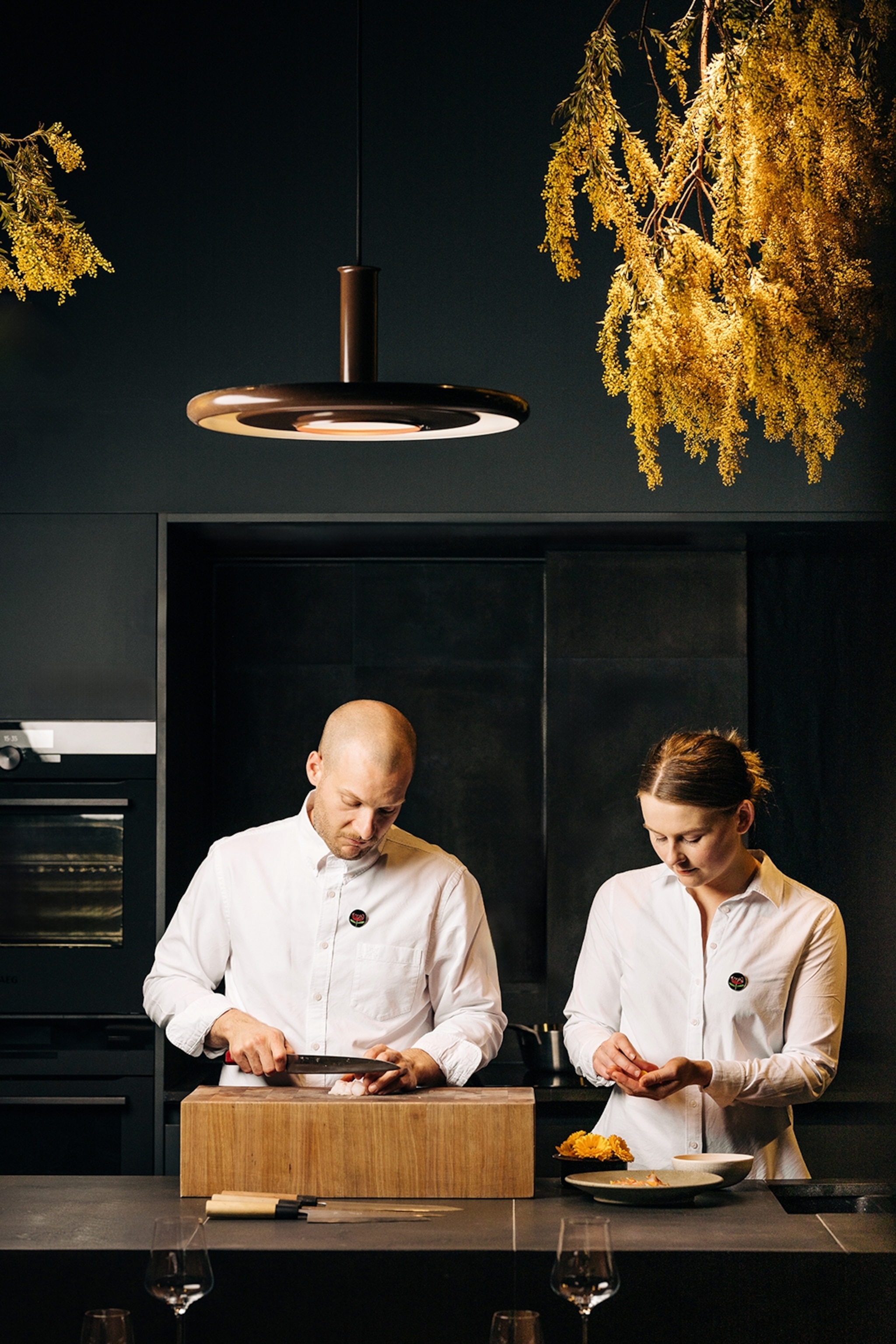 Two chefs, one male and the other female, preparing ingredients in a clean and sophisticated restaurant, dressed in neat chef coats.