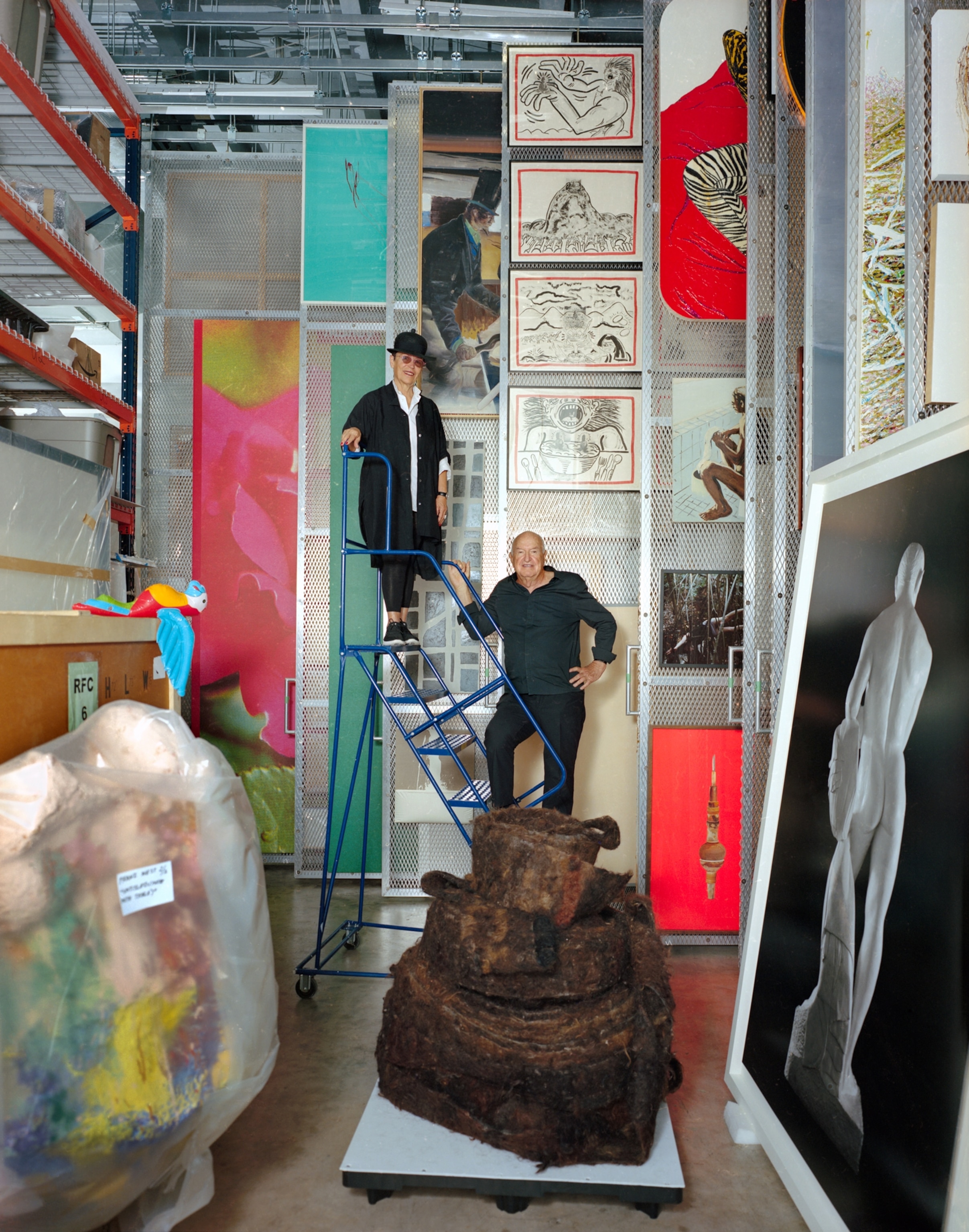 two art collectors standing on a latter surrounded by art in a storage unit