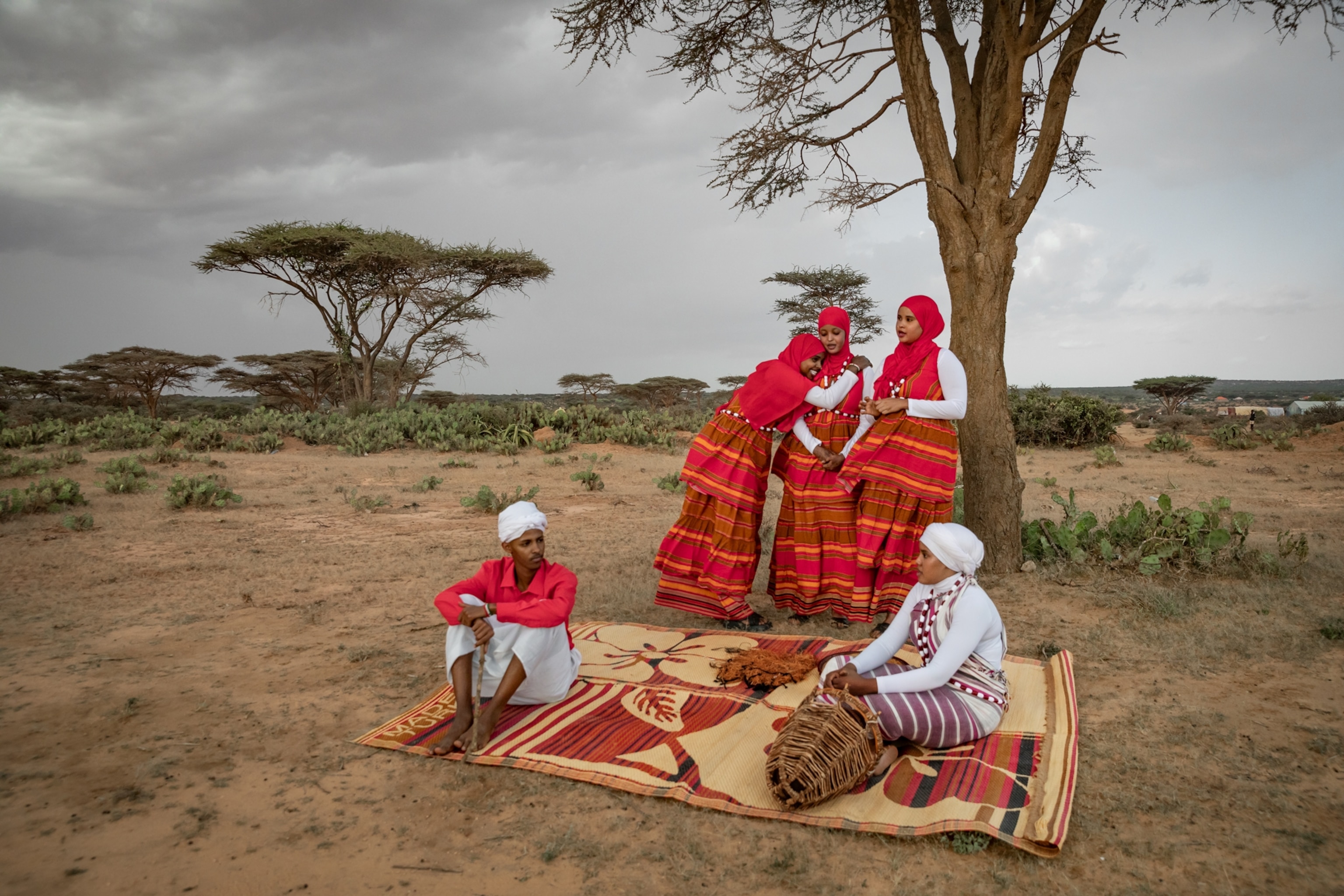 Outside in the drylands three women in bright red and orange dresses laugh and lean on each other as a man and woman sitting below them on a kabad relax.