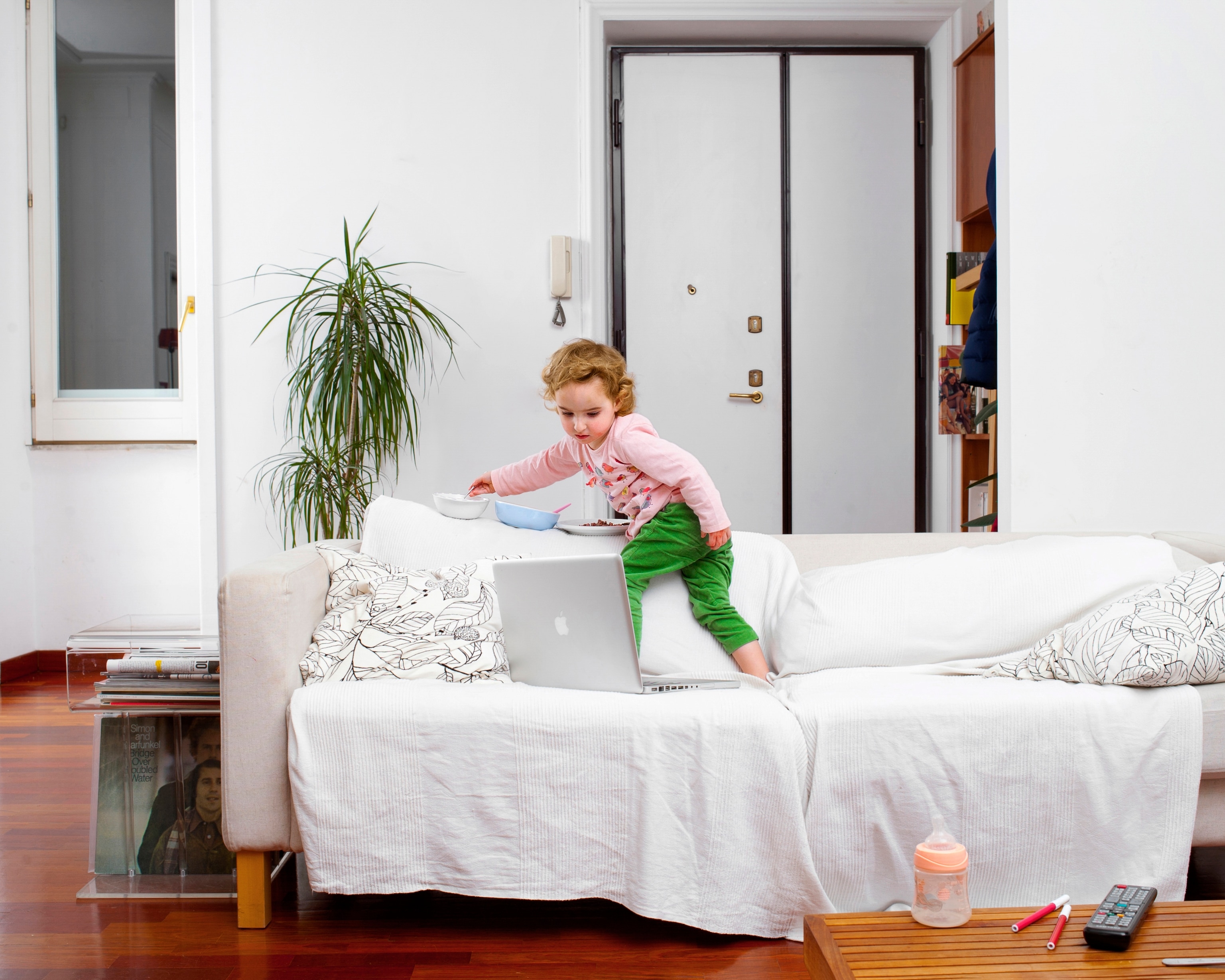 A two-year-old white girl stands on a sofa in a clean apartment. She watches a laptop next to her on the sofa and eats out of bowls lined up on the back of the sofa.