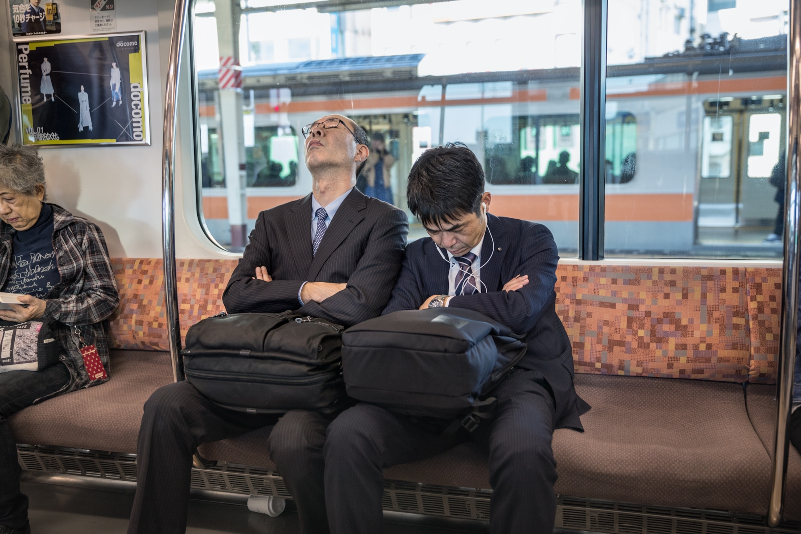 two men sleeping on a subway train with their arms folded