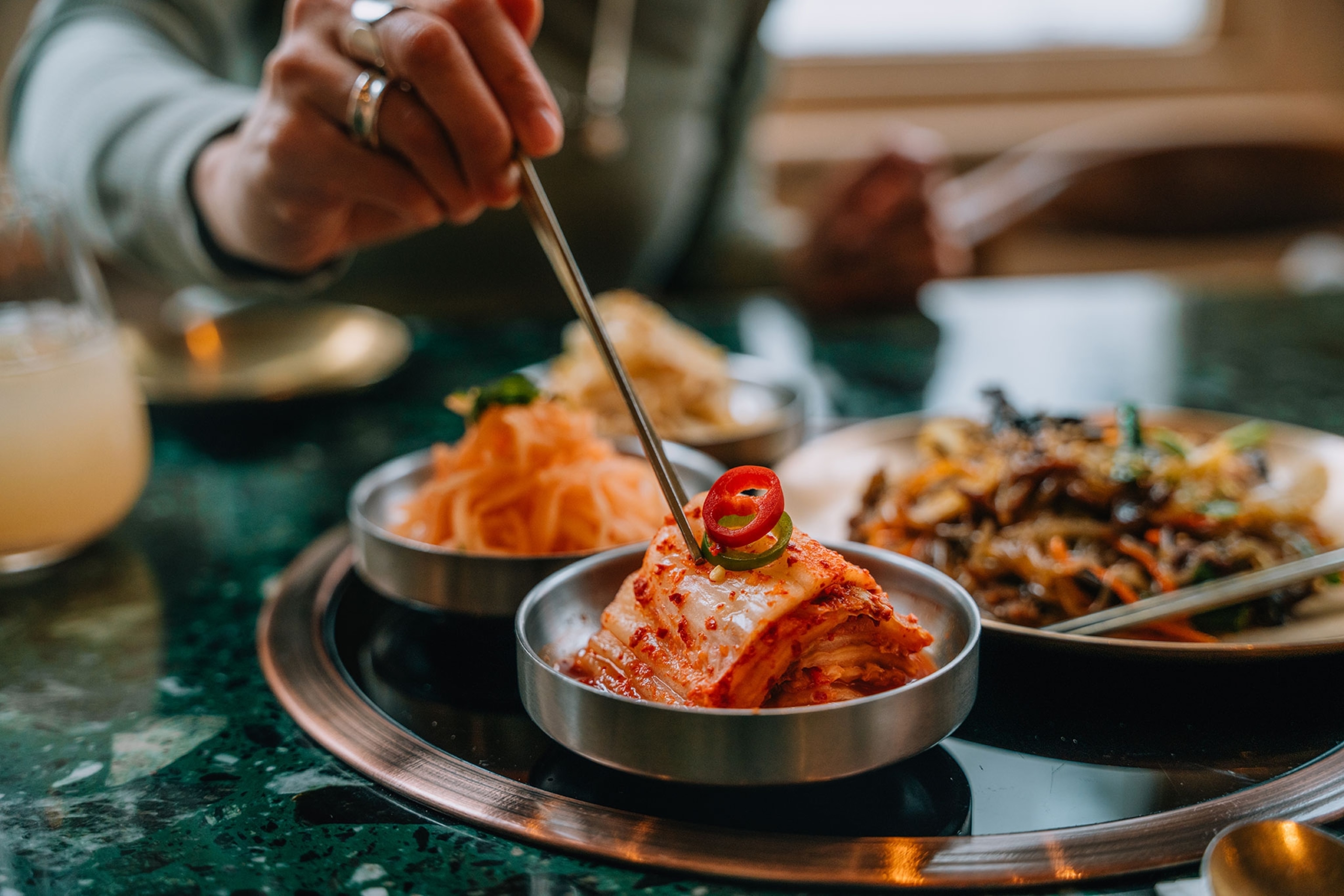 A person's hand reaching over a table of food for a bowl of kimchi with silver chopsticks.