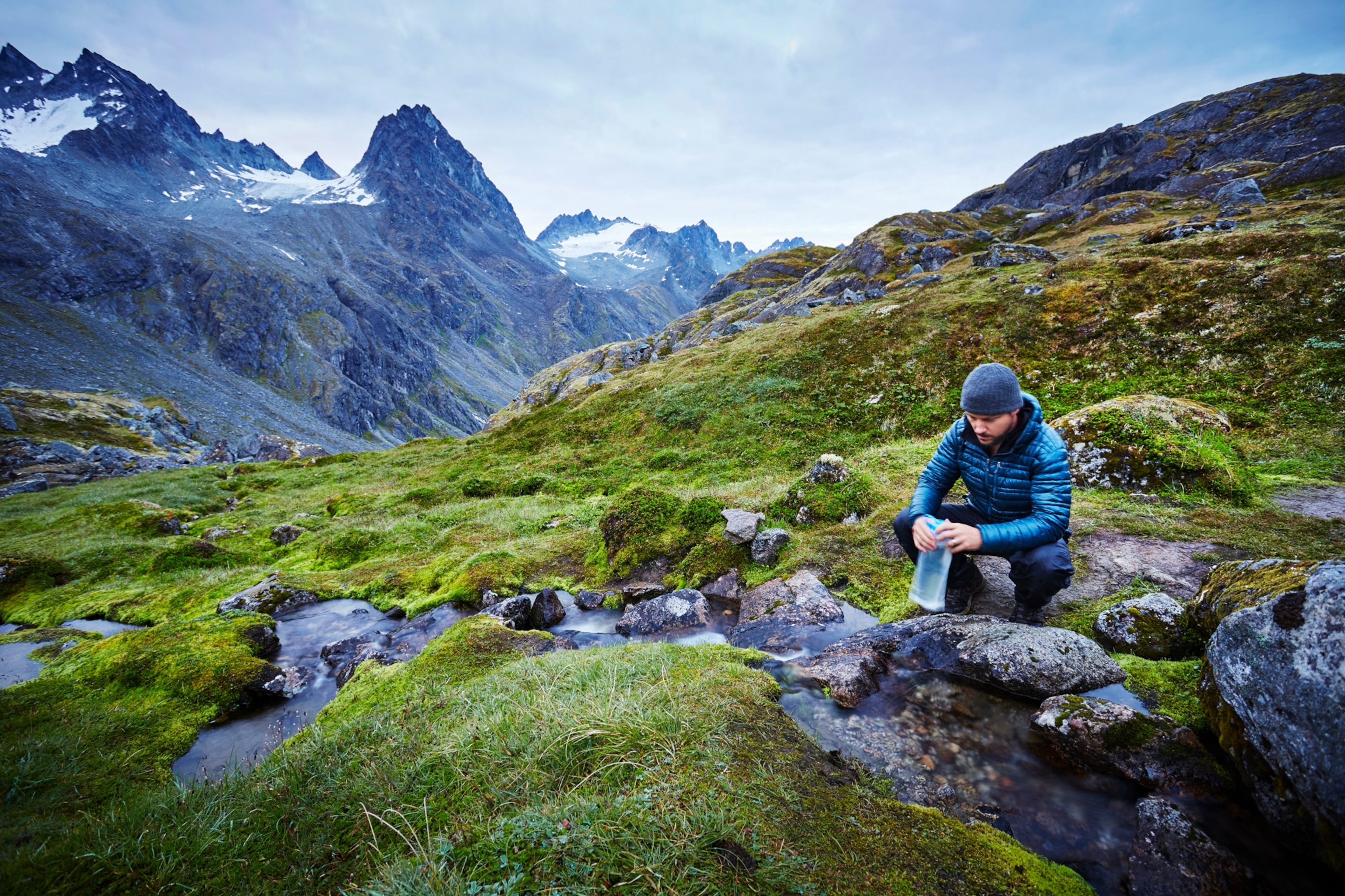 A man bent over by a stream as he fills a water bottle; snow-capped mountains fill the horizon in the background