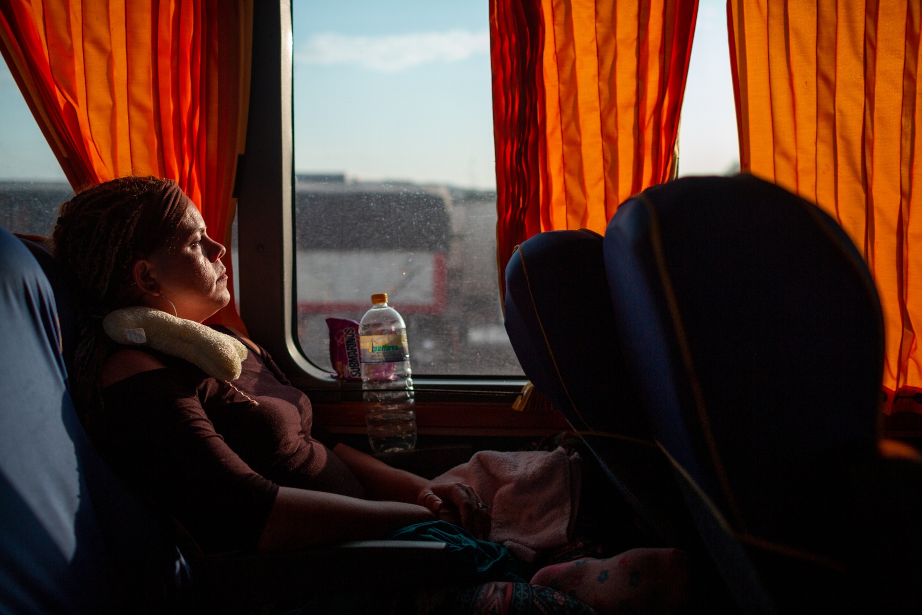 a woman sitting on a bus looking out a window with orange curtains
