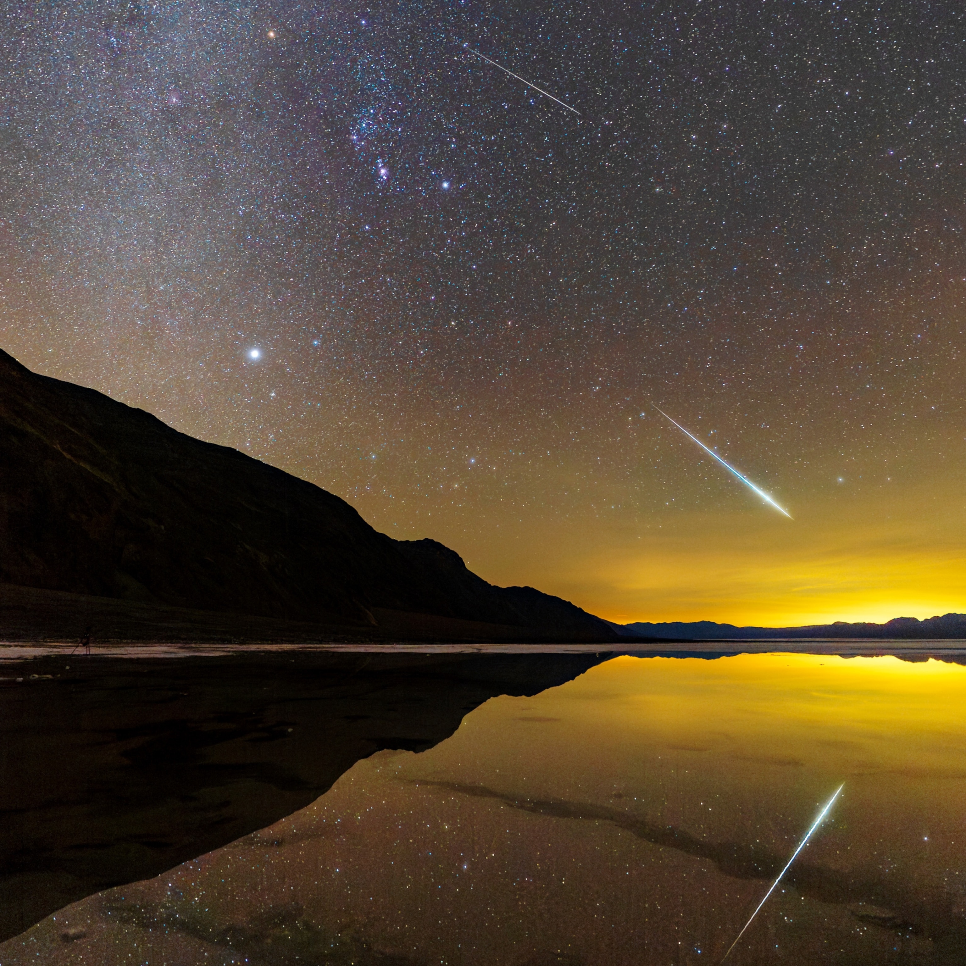 The bright streak of a meteor against a starry sky is reflected in the waters of a lake.
