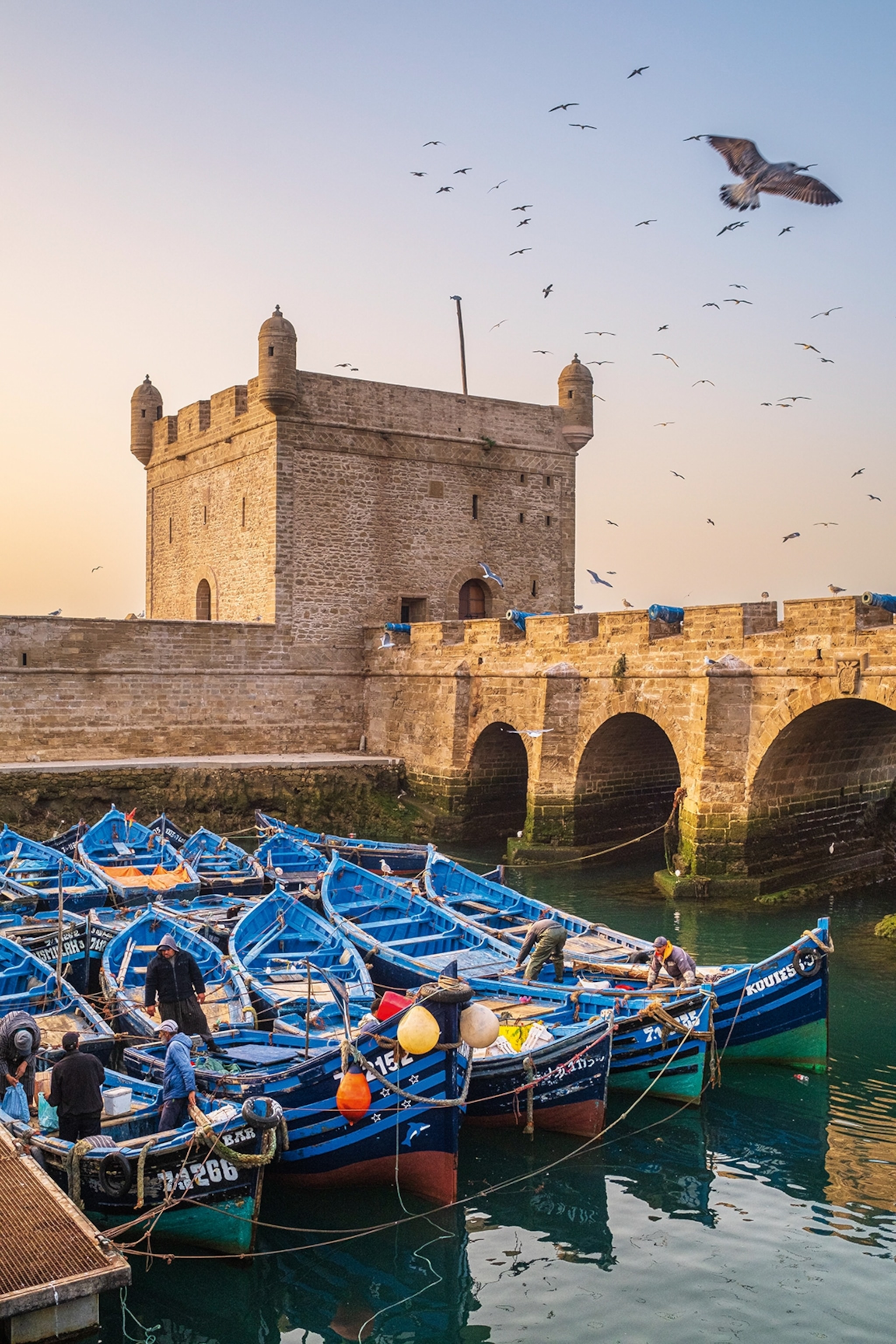Rows of small fishing boats tied to a dock in front of a stone fortress with an arched bridge leading to it.