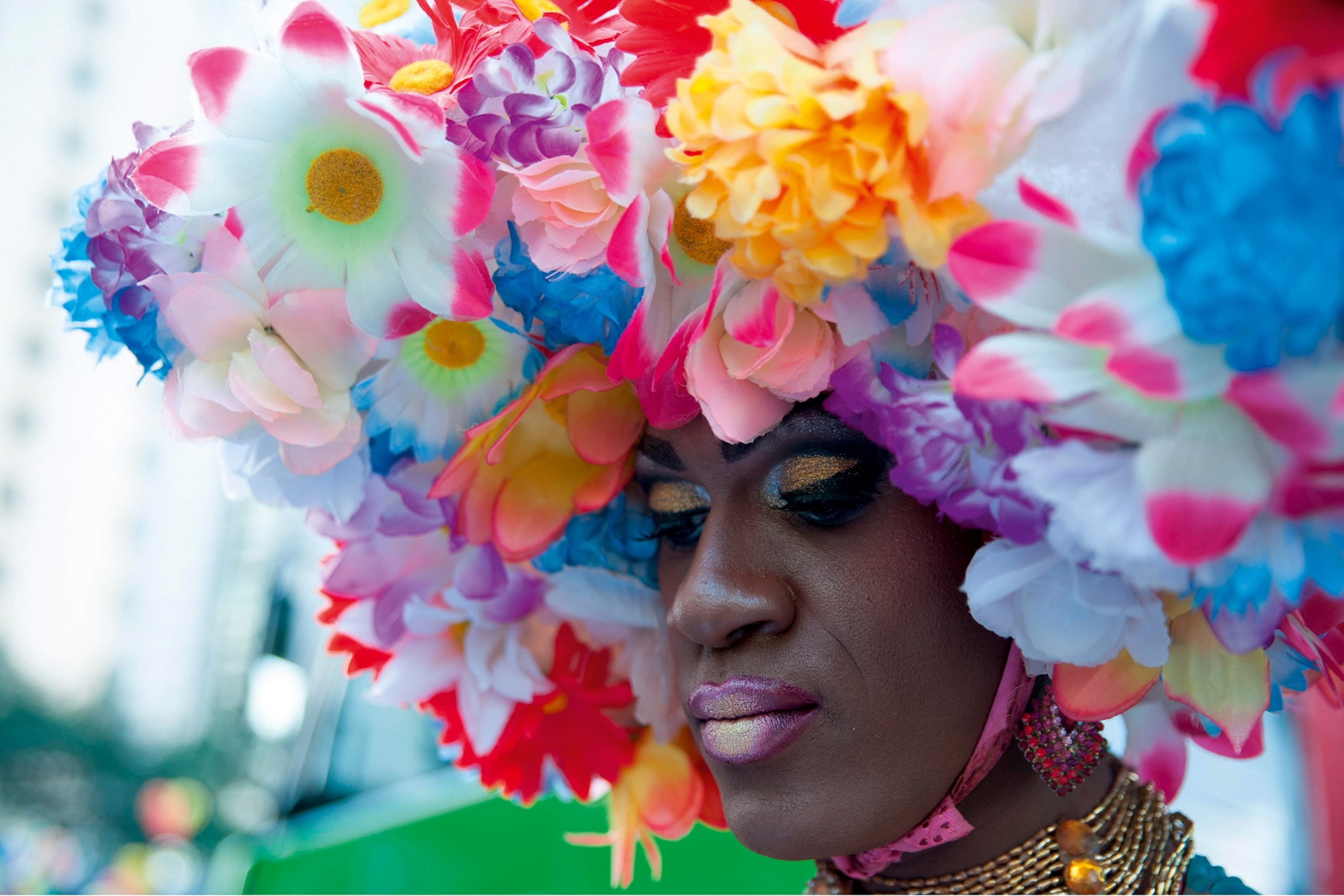 Someone attending a gay pride parade. They have heavy gold eye makeup and flowers covering their head, secured by a pink ribbon tied under their chin.