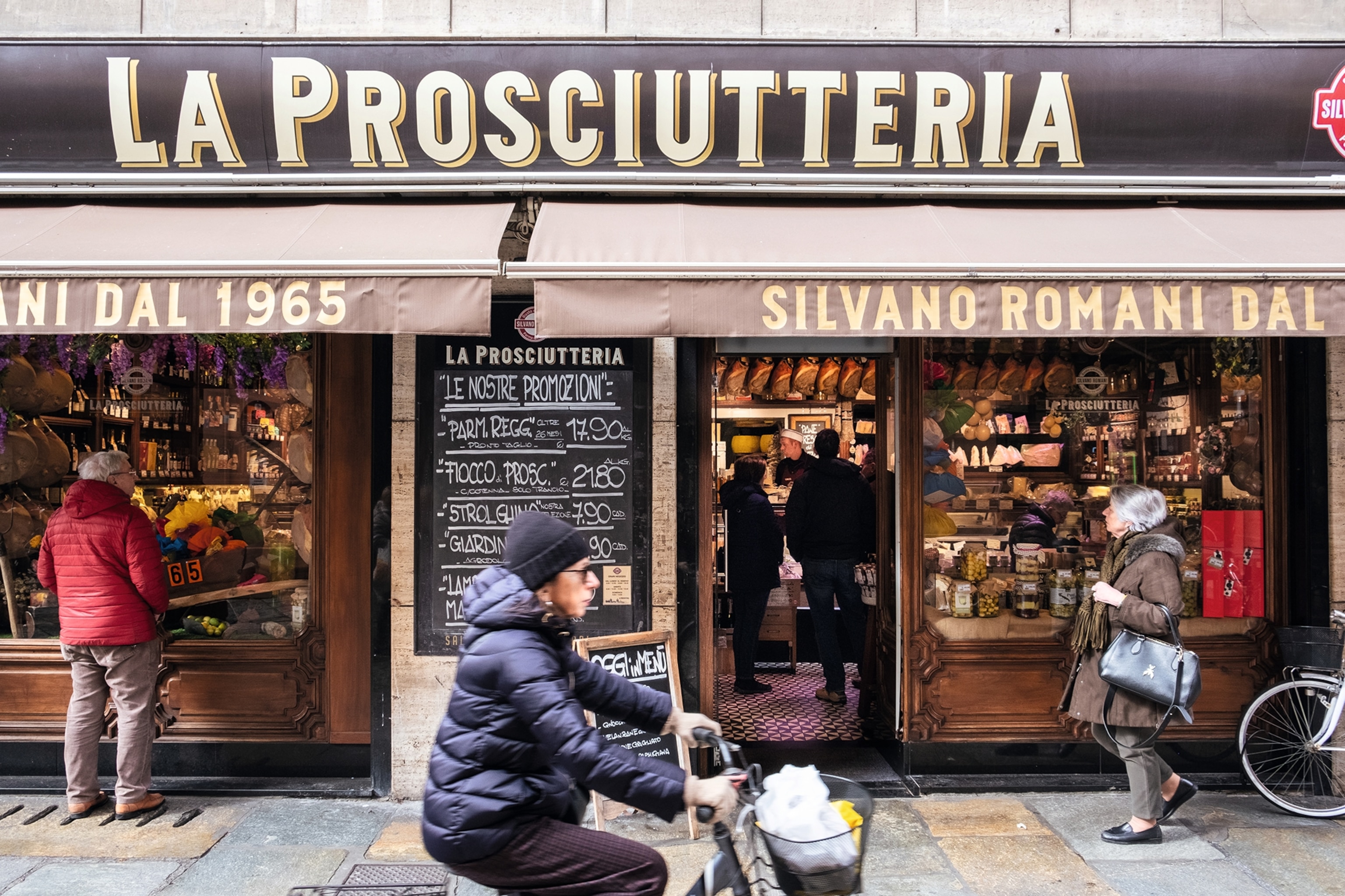 Pedestrians walking past the exterior of La Prosciuttera, with windows on via Farini.