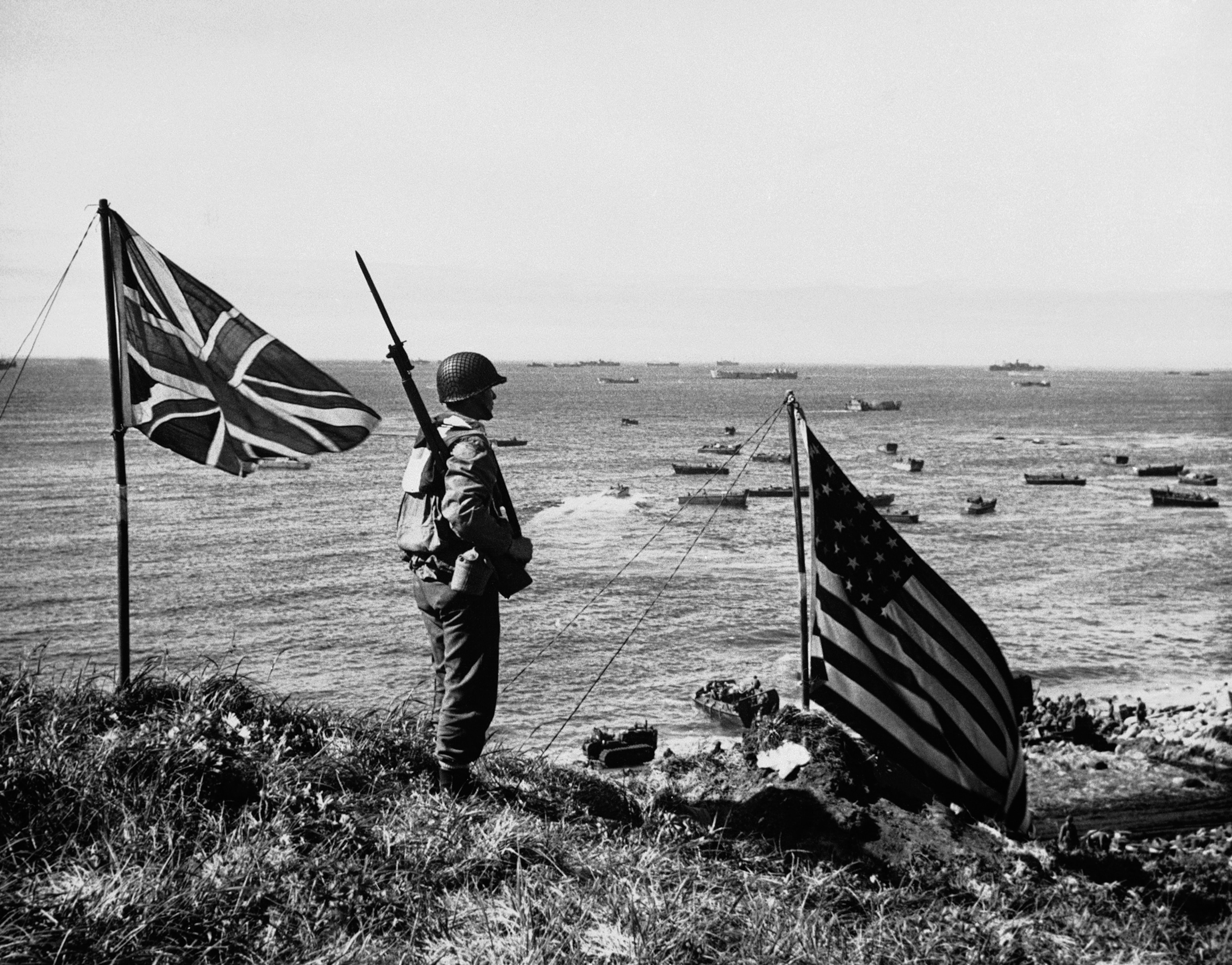 American and Canadian flags fly on top of a hill whilst a US soldier looks out over dozens of ships offshore.