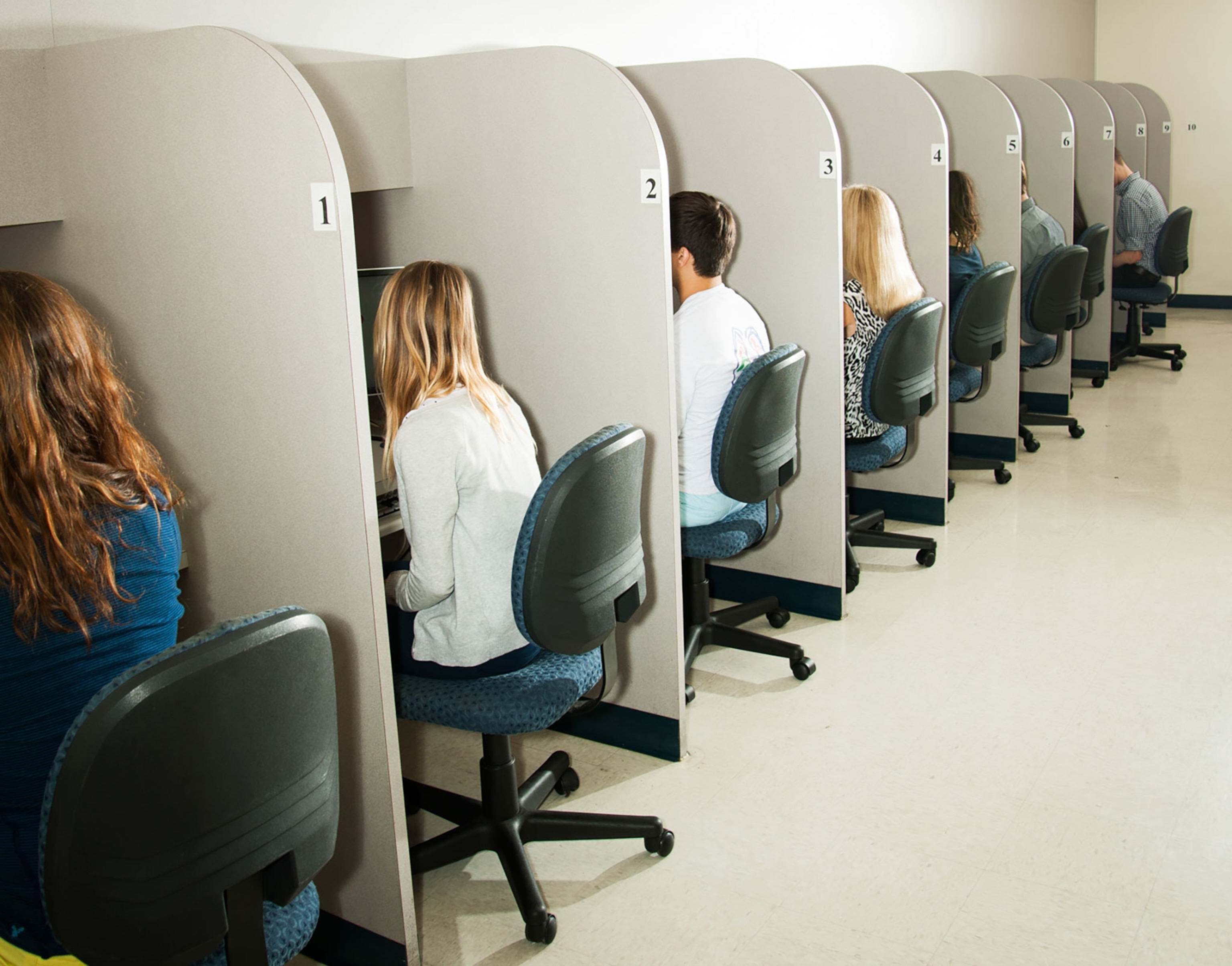 A line of people, sitting in chairs at desks with white partitions.