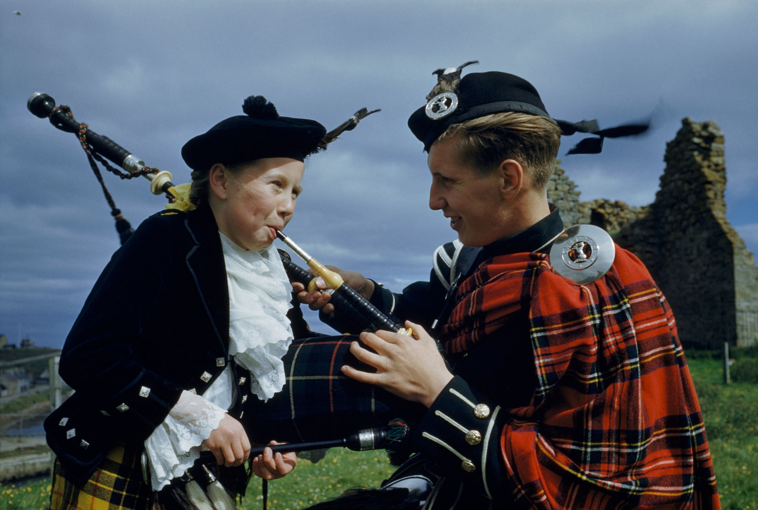 A piper holds a bagpipe mouth piece for someone. Both wear tartans.