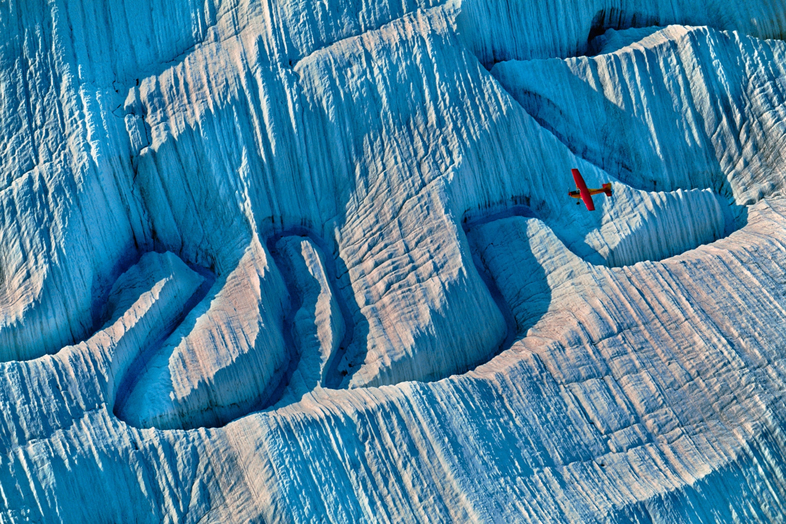 A winding valley of ice below as a red plane flies above contrasting the landscape.