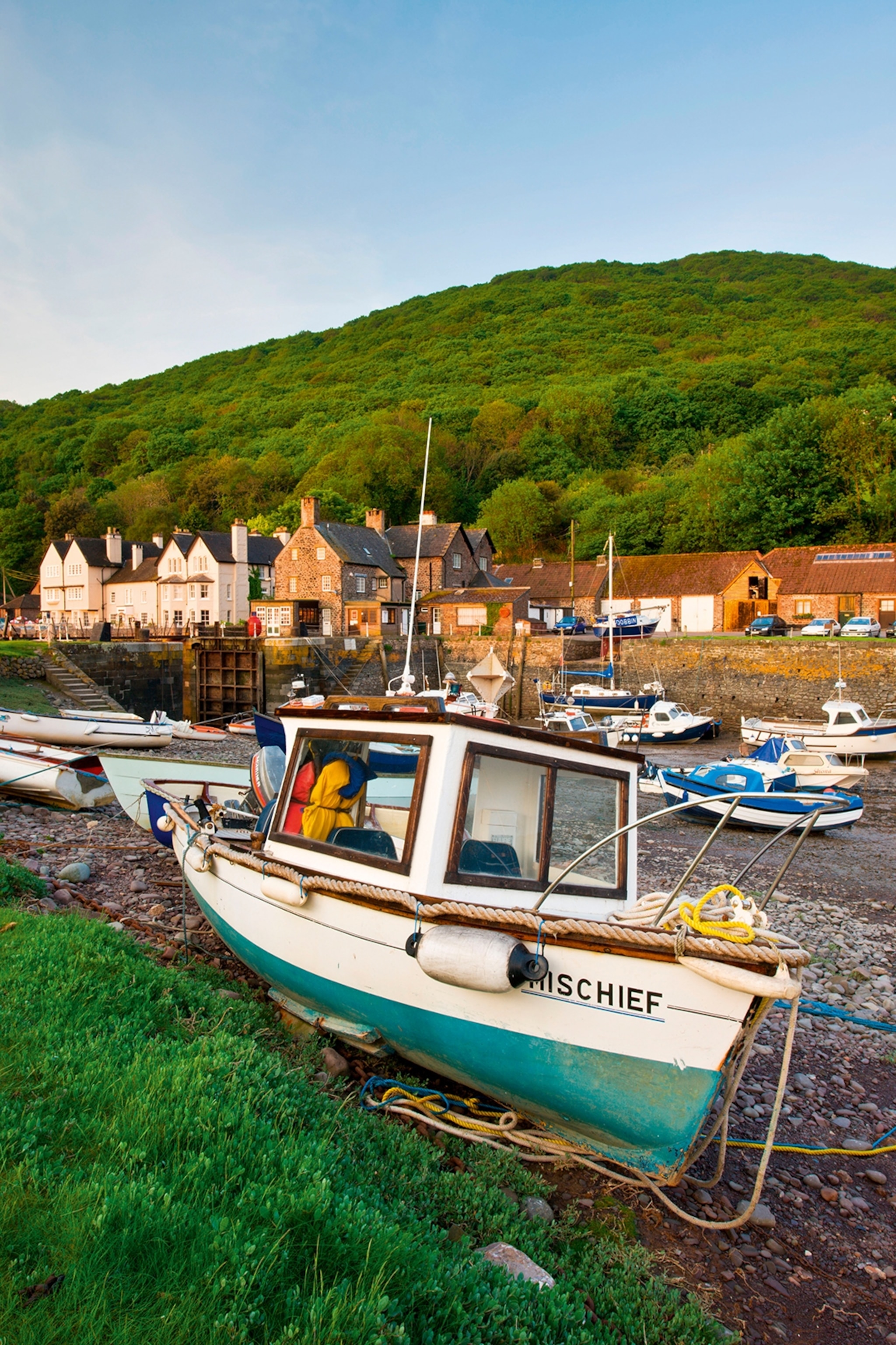 A small fishing boat stuck on concrete during the low tide in a small, British countryside harbor with green hills in the back.