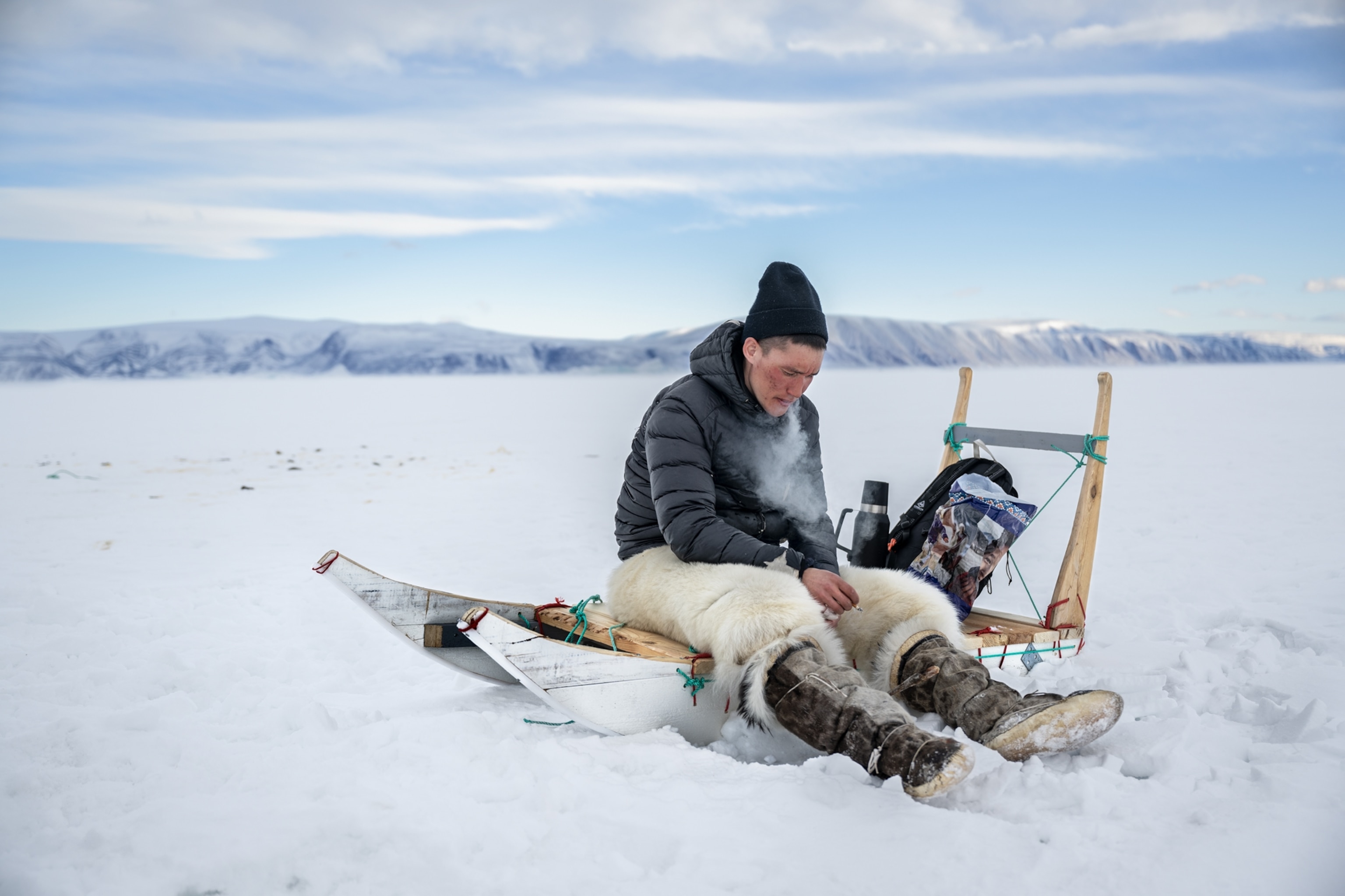 Kvist sits on his sled in the snow, smoking a cigarette. He is wearing white fur bottoms, tall boots, and a black puffer coat.