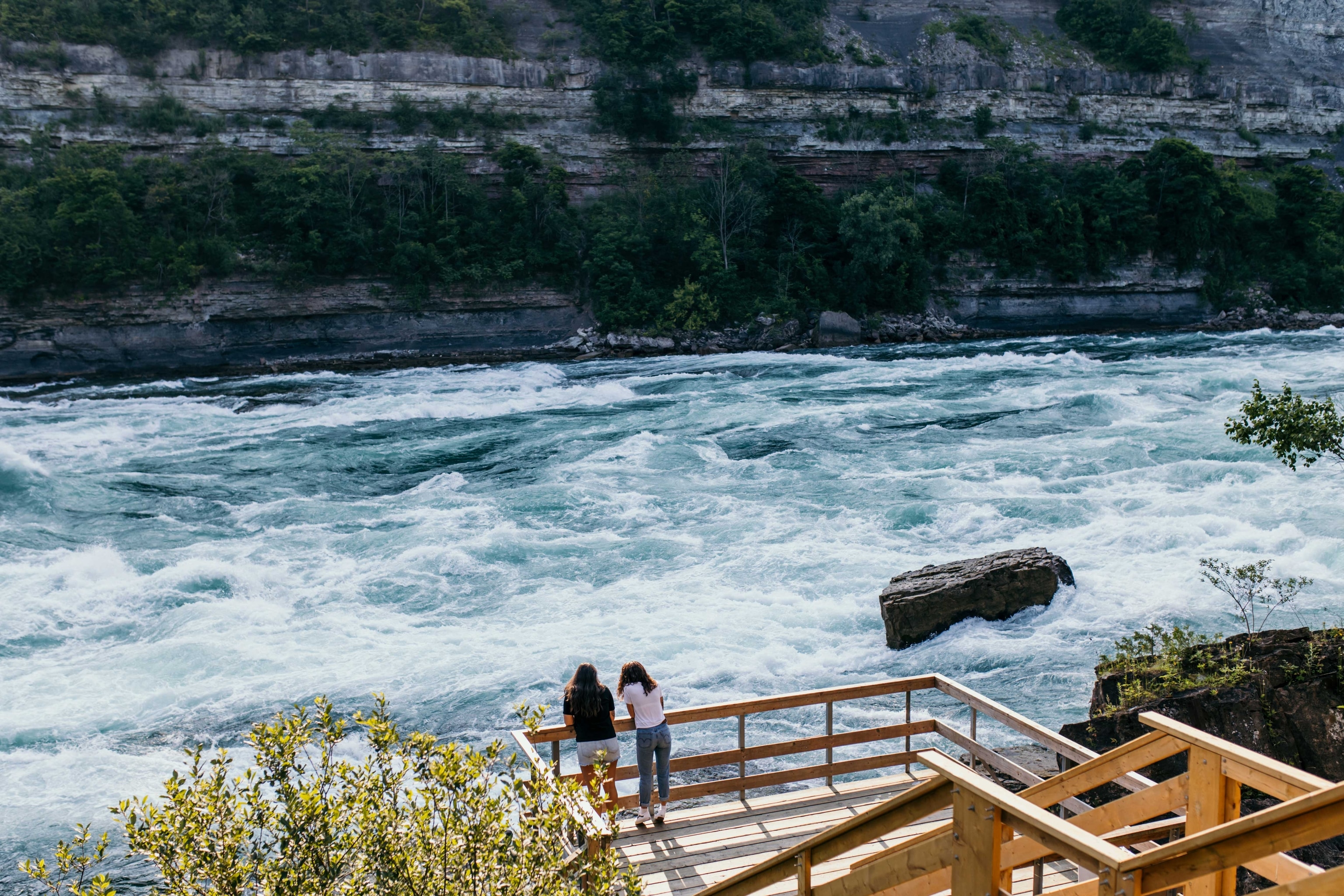 Two women stand on a boardwalk looking at rushing white water