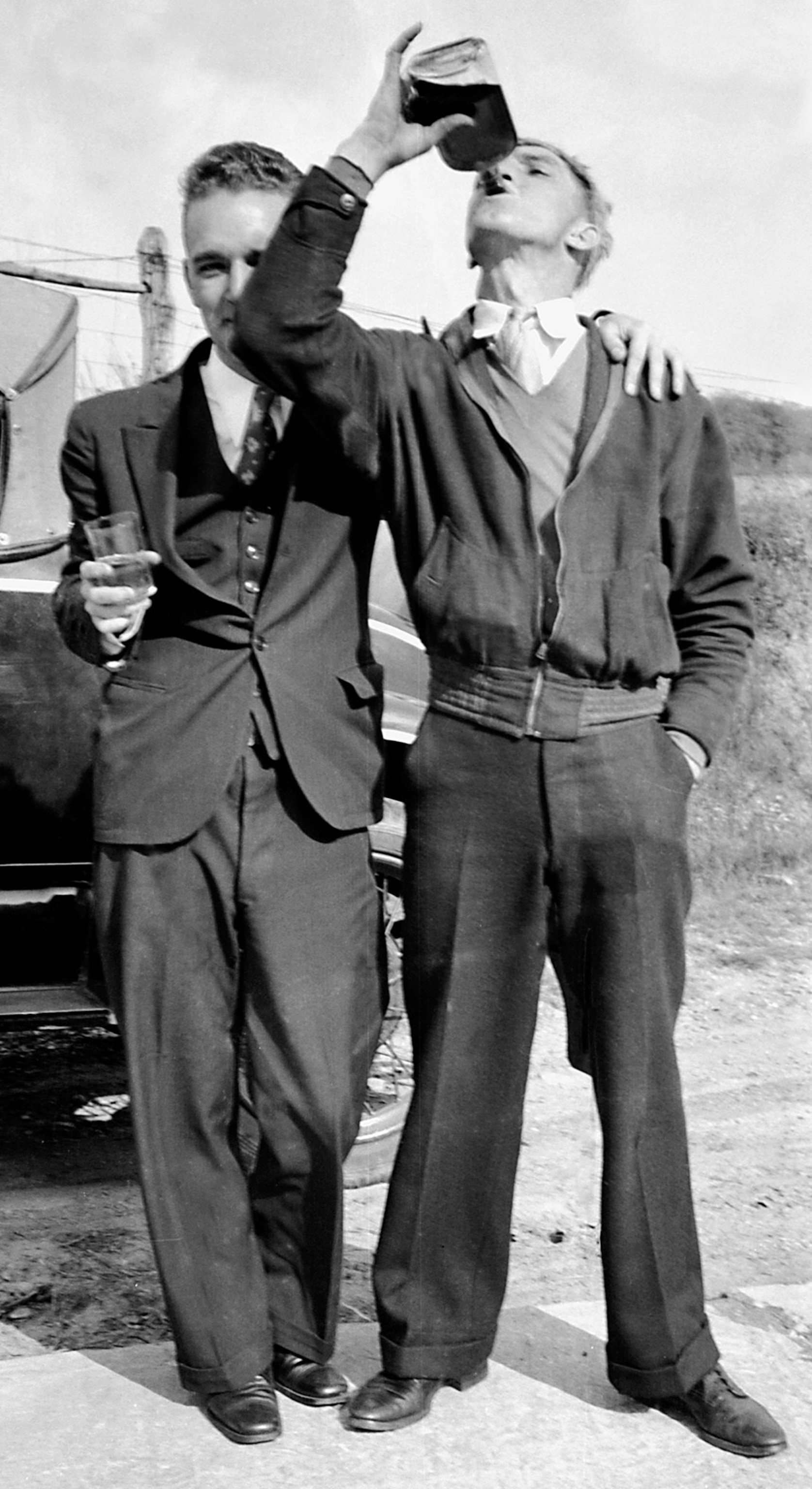 Two man standing one with a glass of liquid facing the camera another with a bottle and head titled back drinking.