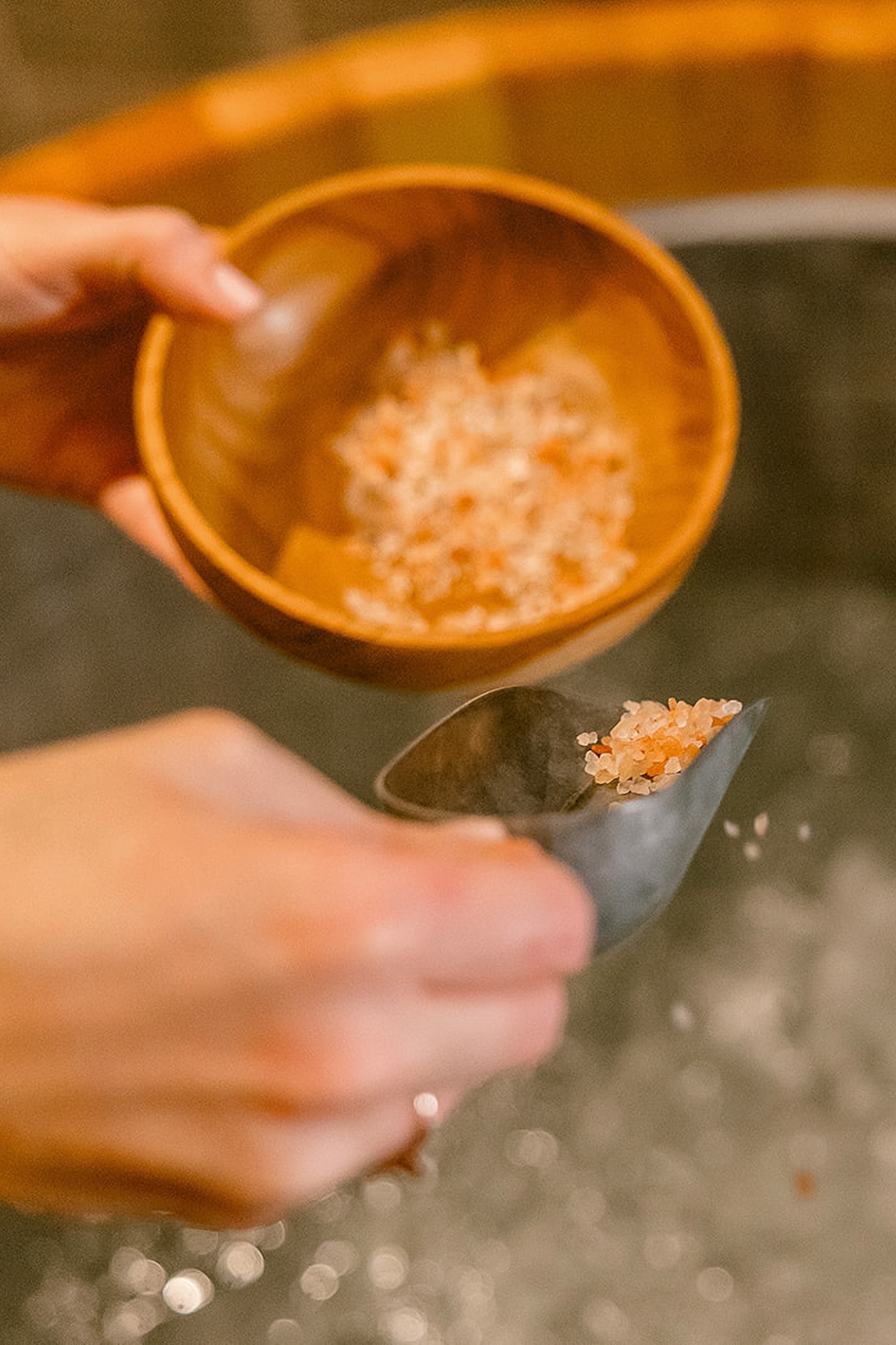 Female hands scooping bath salts out of a wooden bowl into a bubbling bath.