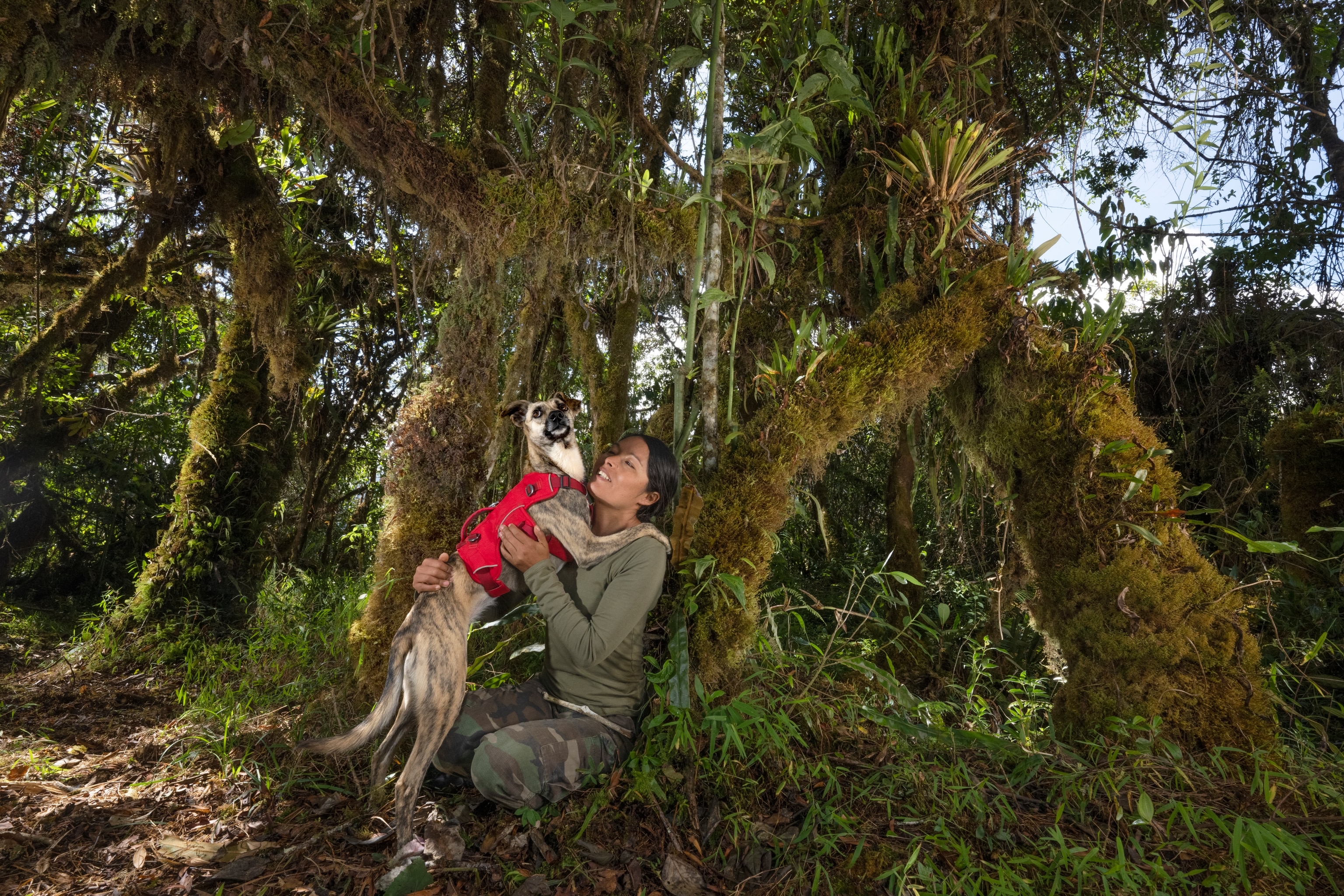 Young woman in camouflage green clothes hugging dog in red vest.