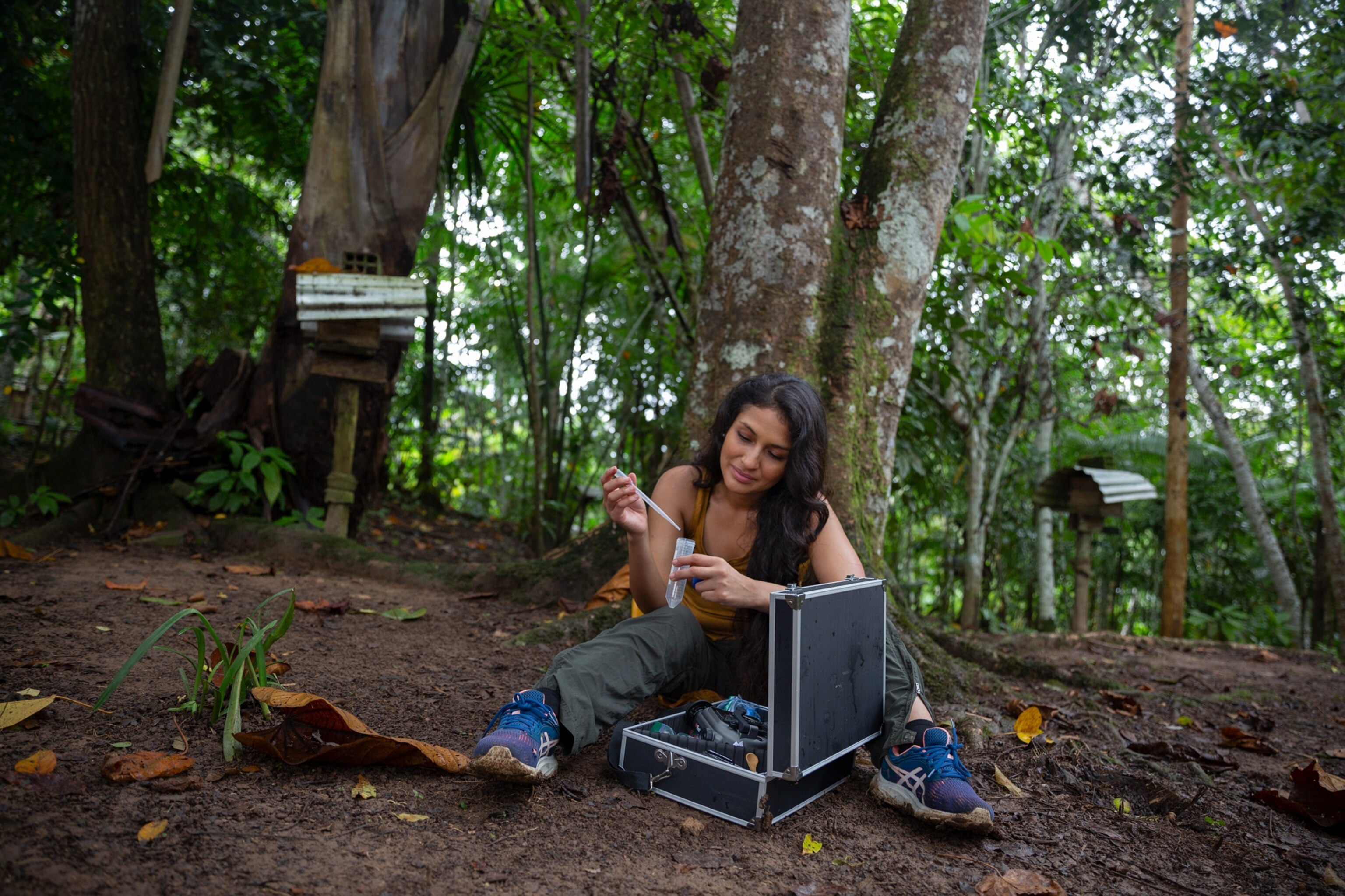 Picture of woman sitting on the ground under forest trees with lab case and testing tubes.