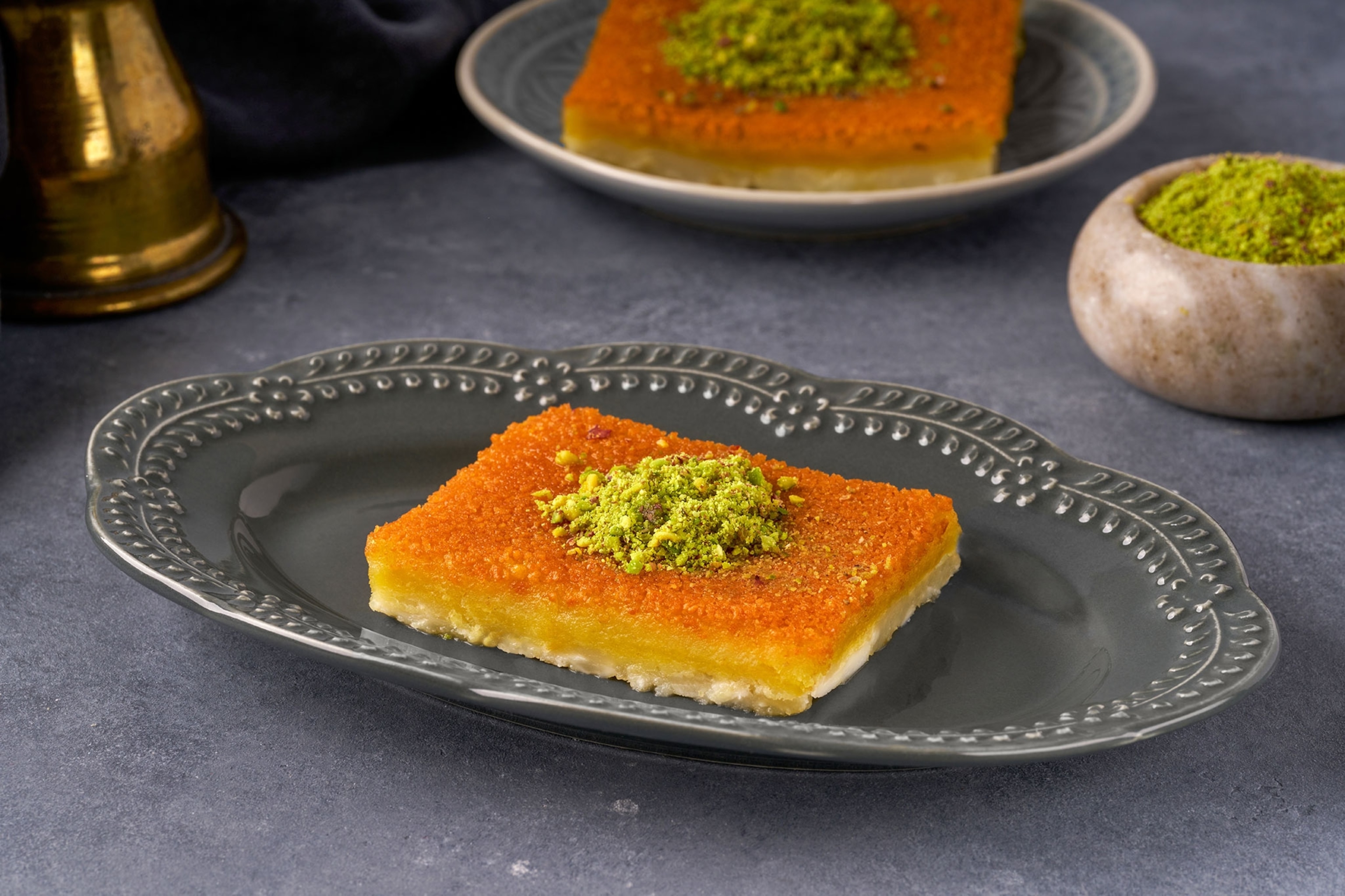 A fried pastry with crushed pistachios on top sits on a grey plate, with another portion and another bowl of crushed pistachios in the background.