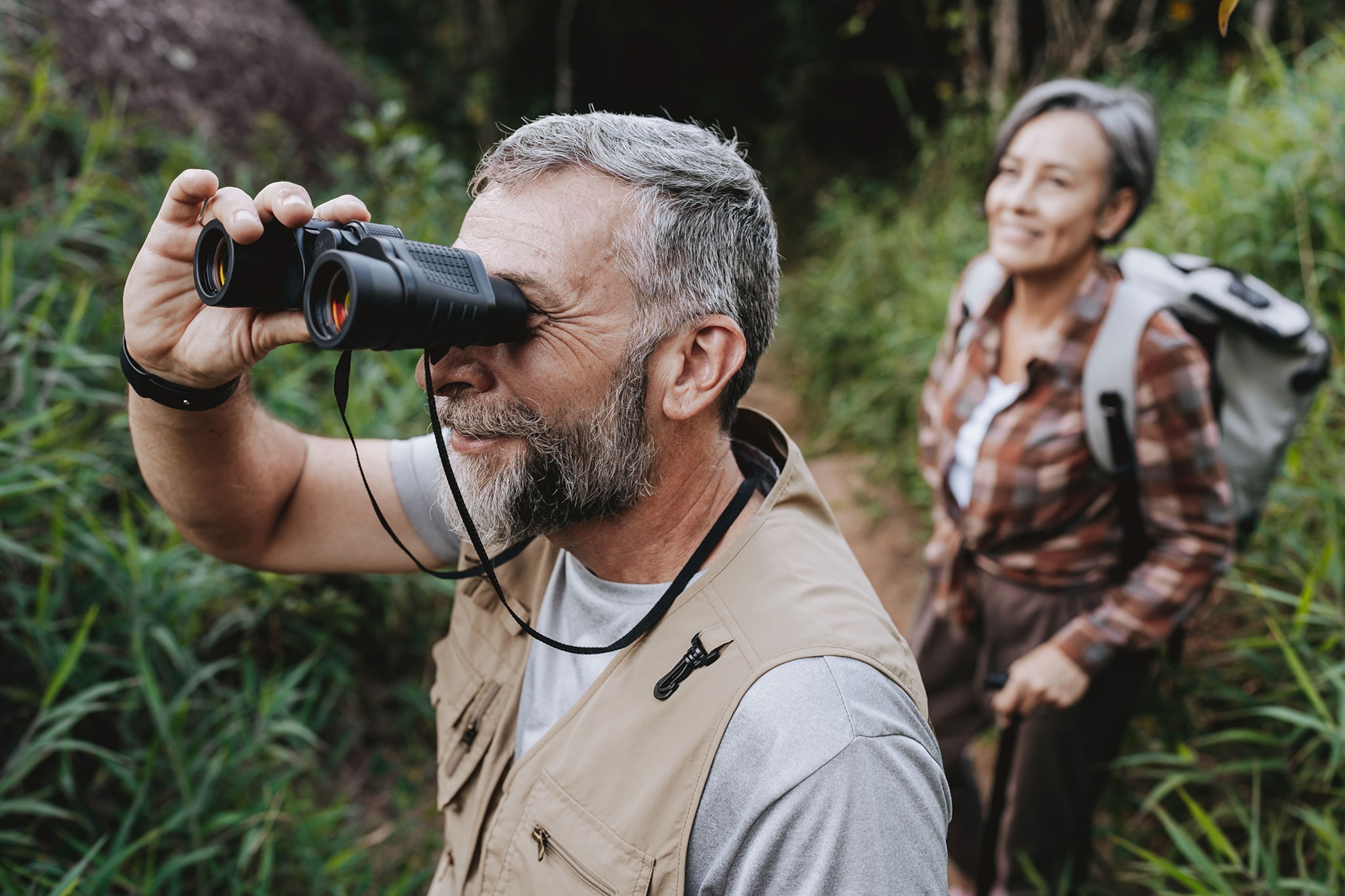 Older man obsering nature with binoculars as woman stands behind watching.