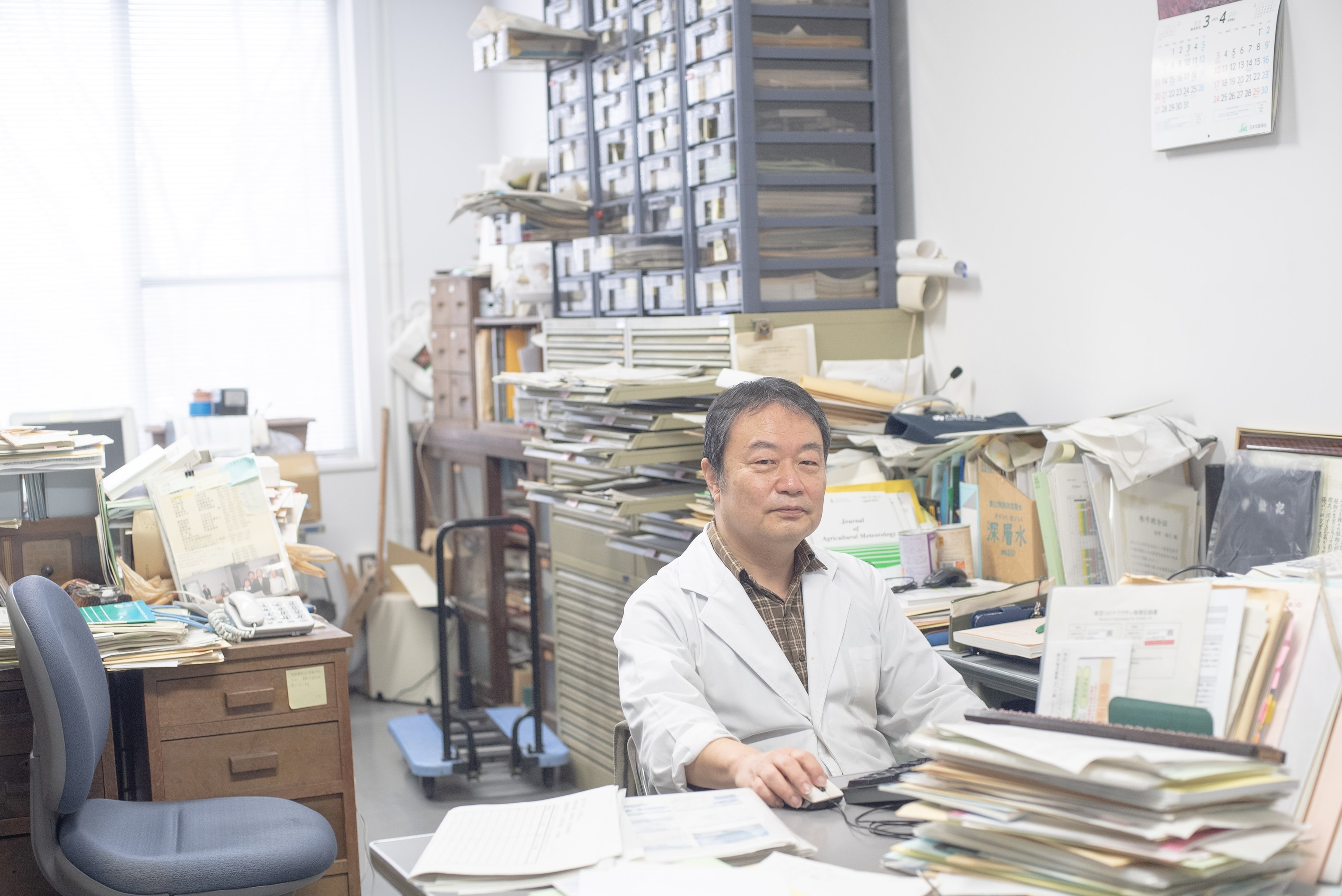 A researcher wearing a white jacket sits at his desk surrounded by books and papers.