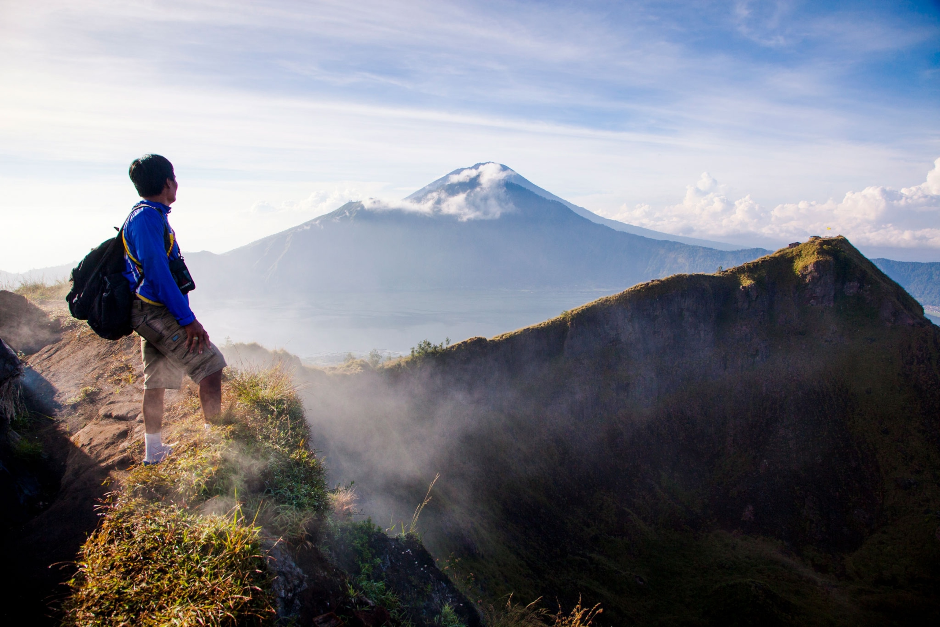 a hiker on a tall peak looking out at two additional peaks with a layer of clouds blocking the bottom of the mountain