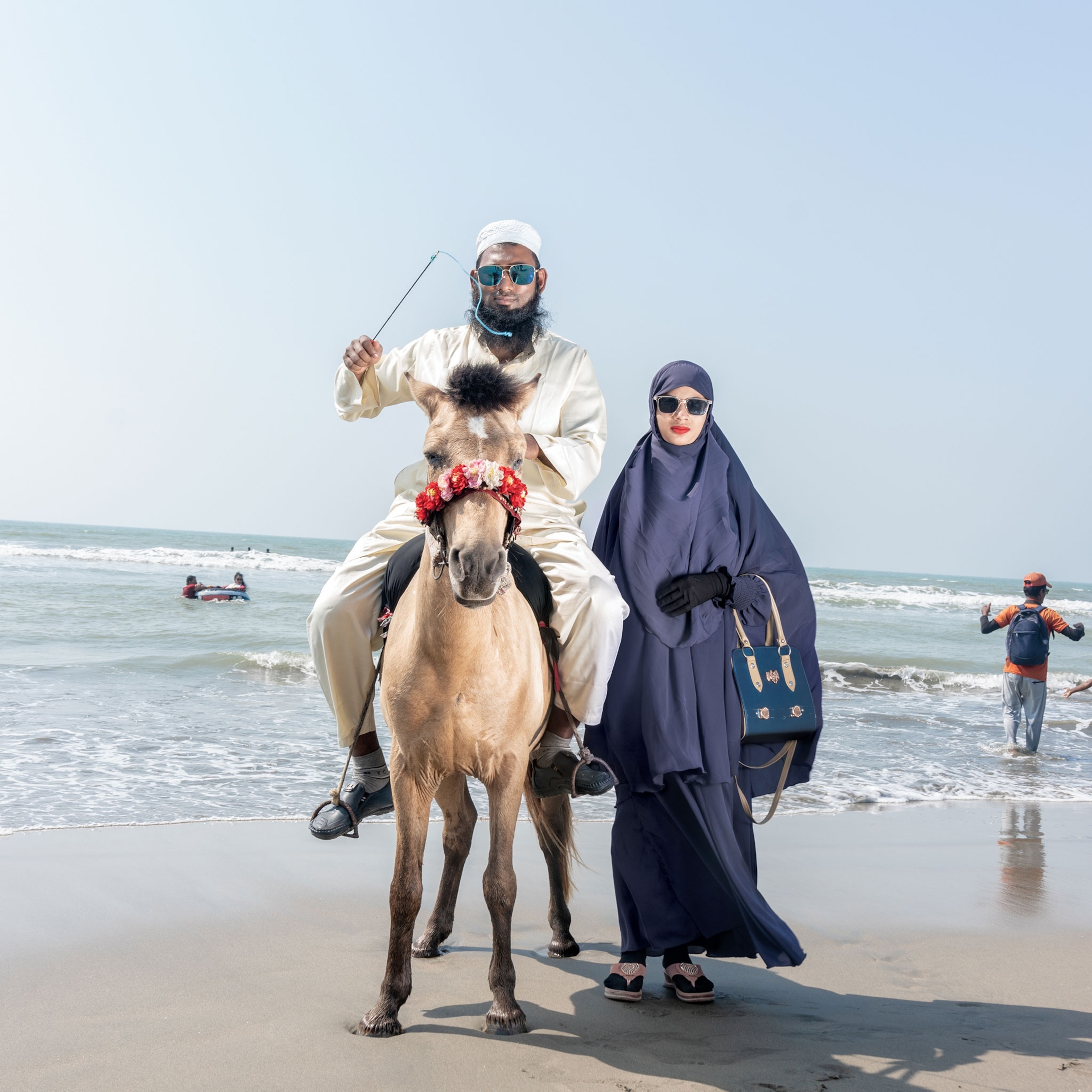 Picture of man in traditional white clothes and sunglasses sitting on hourback and woman by his site in traditional cover and sunglasses wearing bright red lipstick.