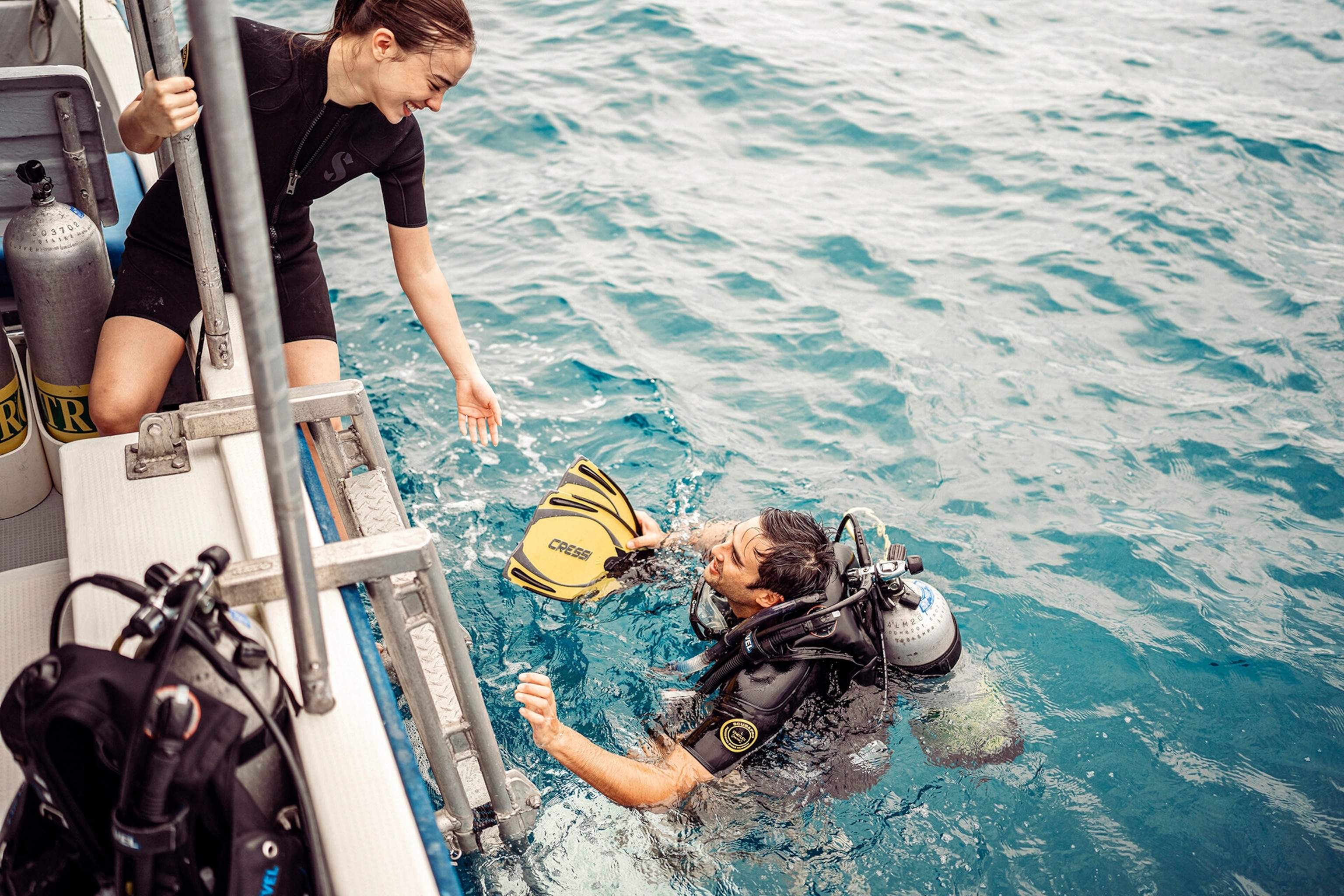 A female diver on a boat reaching out a helping hand to a male diver in the water.