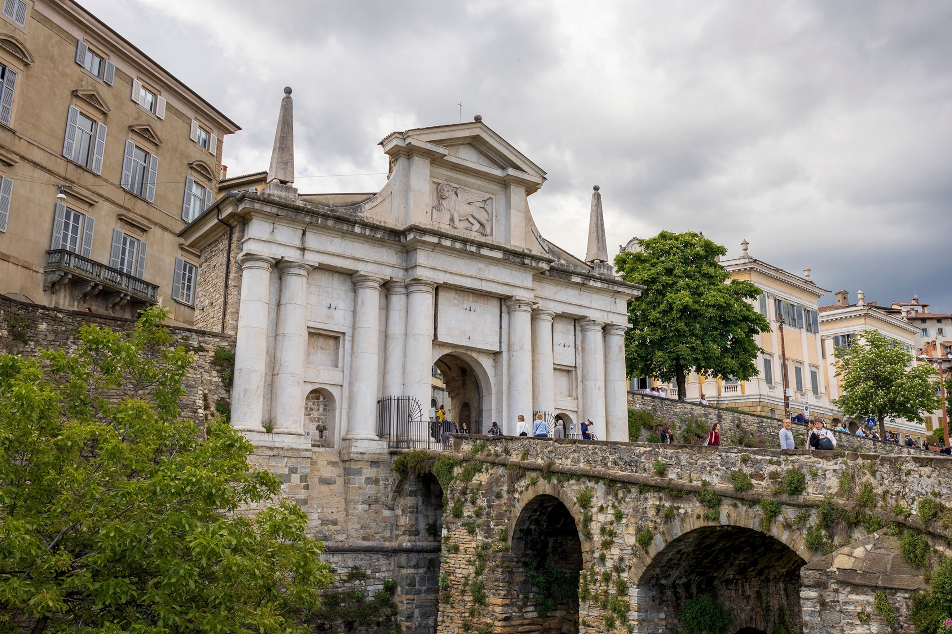 A large white marble entrance into the city behind it with a stone bridge covered in vines coming out from the entrance on the right side of the frame.