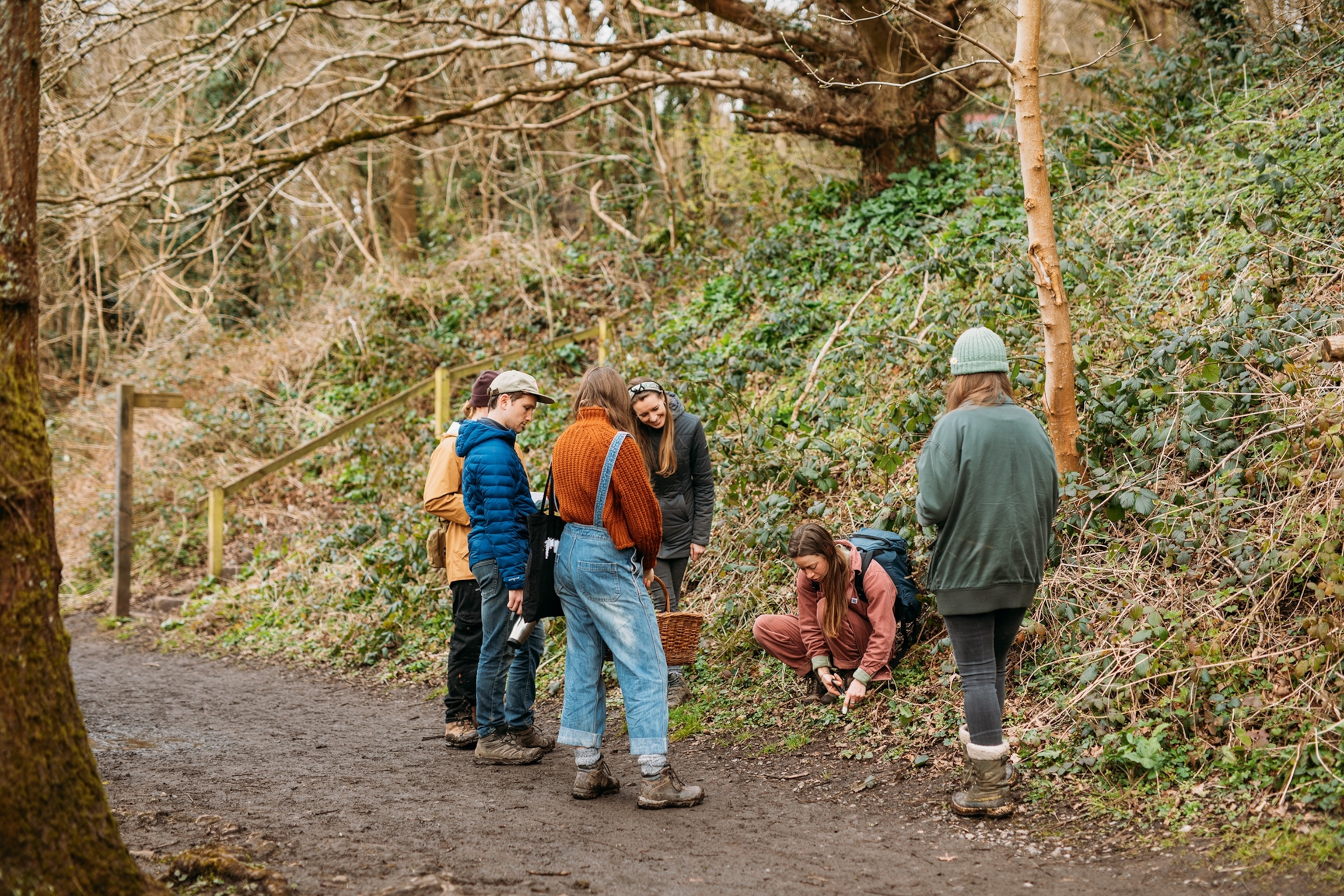 A group of young people in a wet forest, arranged in a circle around a tour guide presenting herbs on the ground.