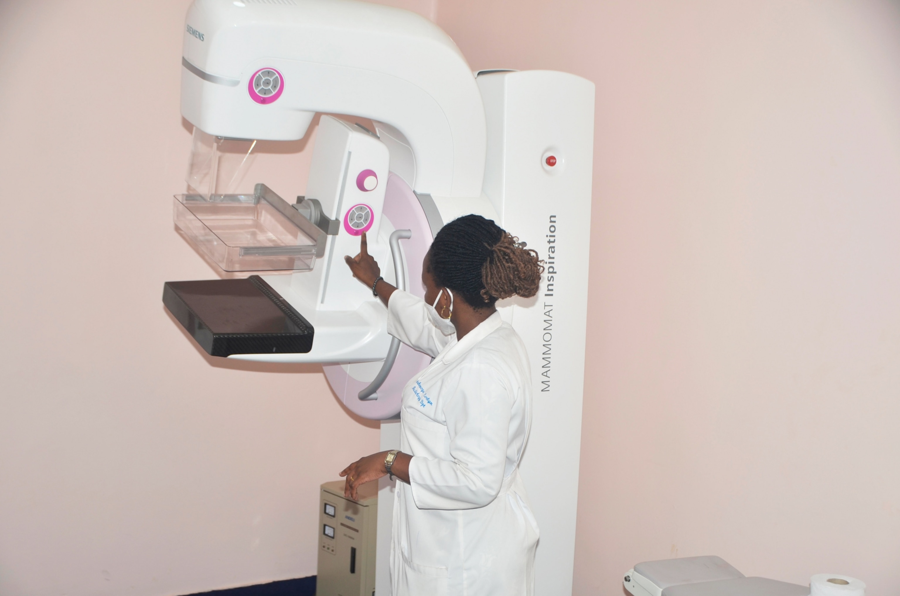 A nurse points to part of a large white mammography machine, which is standing in an examination room with light pink walls.