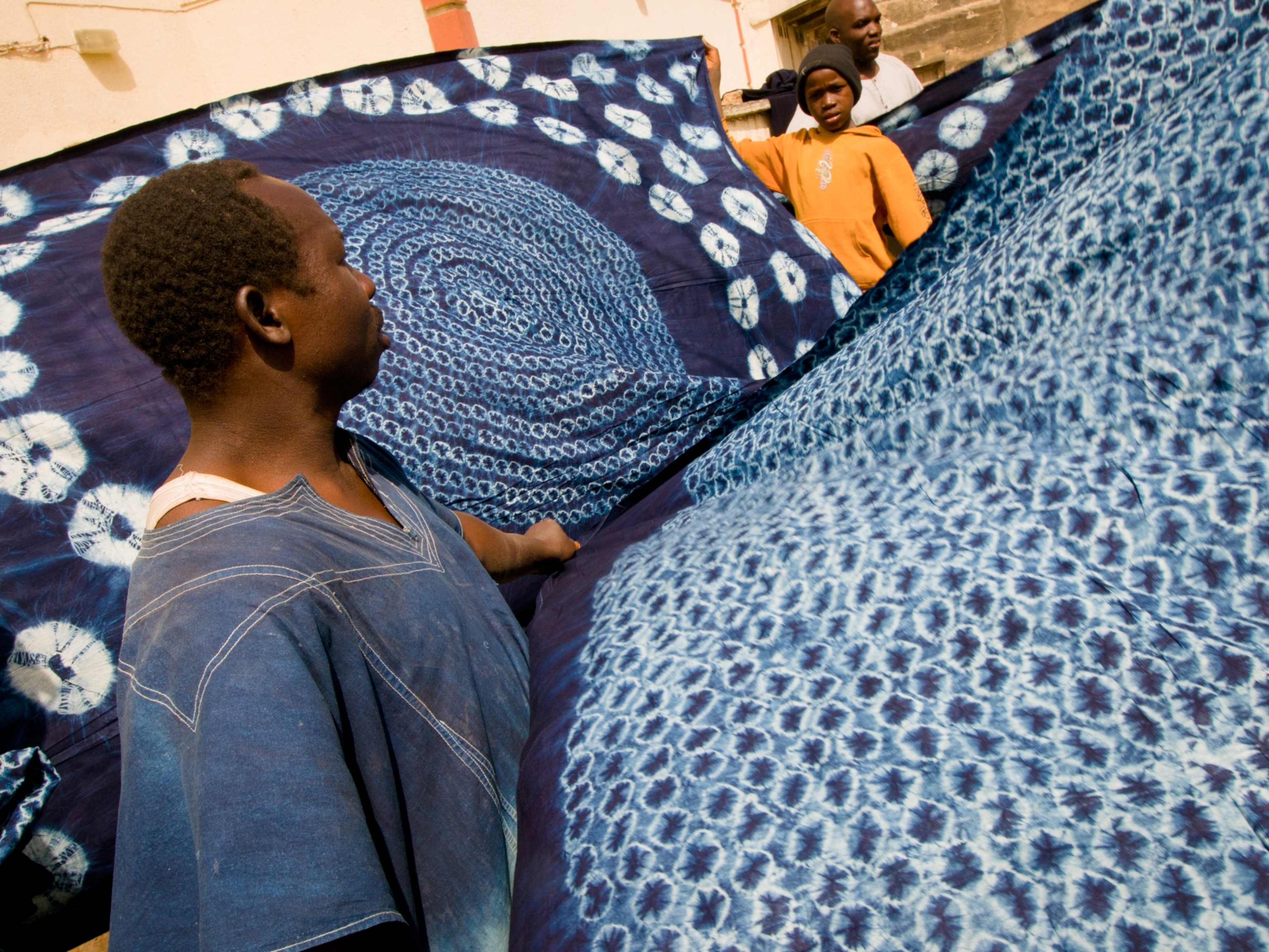 Men hold up large textiles they have died a deep indigo, leaving intricate circular patterns of un-dyed white cloth resulting from their tie-dye technique.