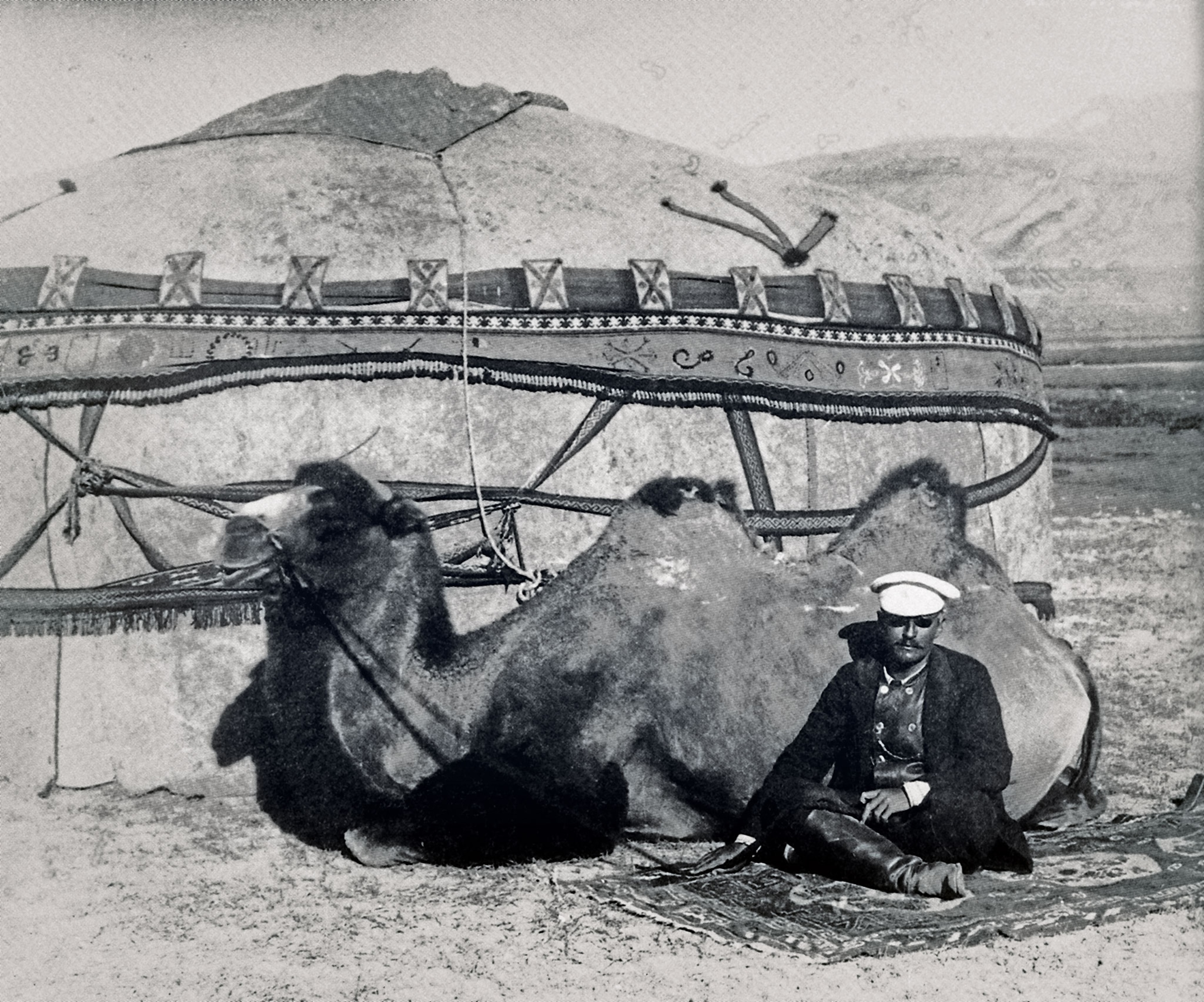 A man sits on the ground next to a sitting camel in front of a structure in a black and white photo