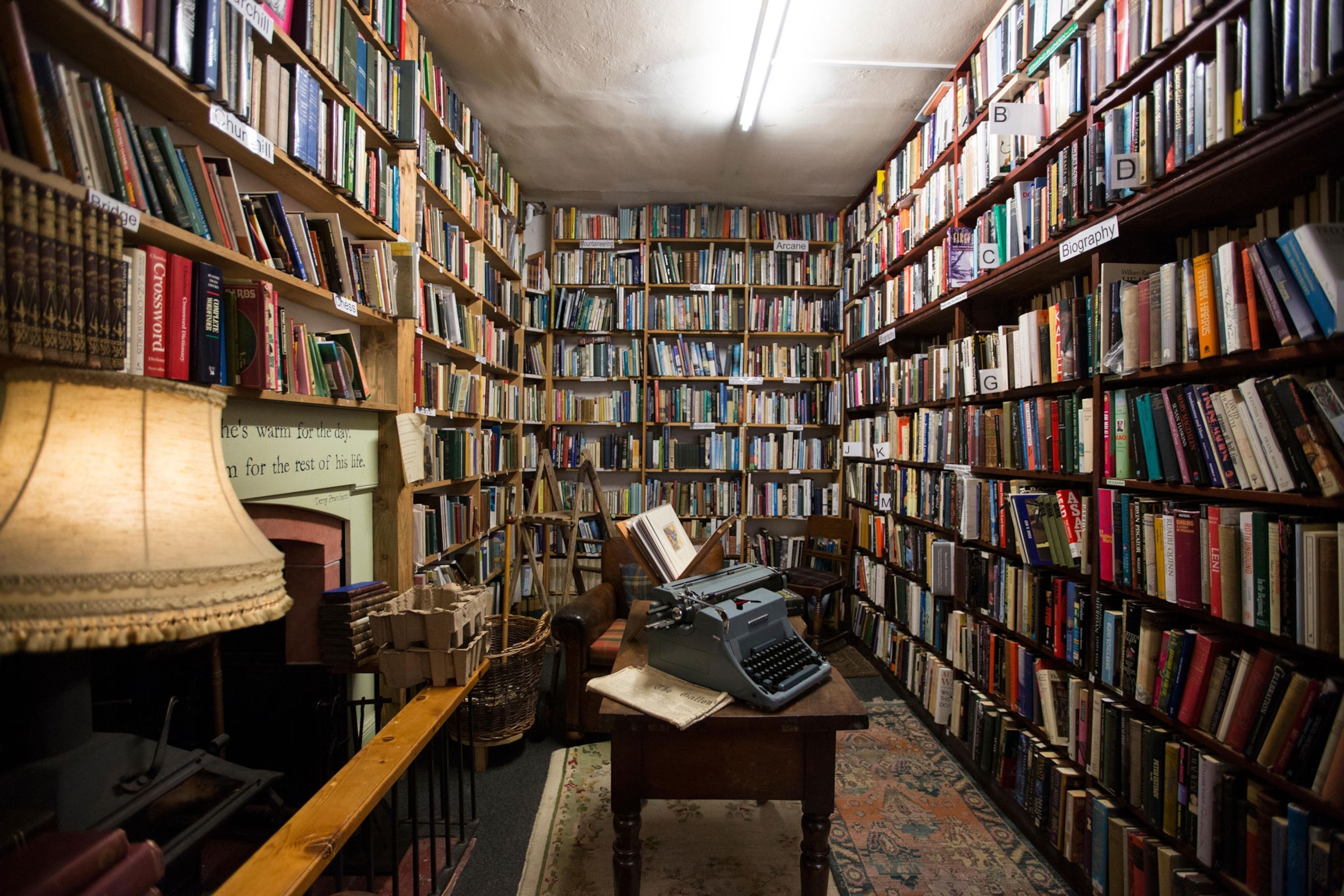A picture with wall to wall books, a lamp, a fireplace, and a vintage typewriter on a vintage desk in the middle of the room.