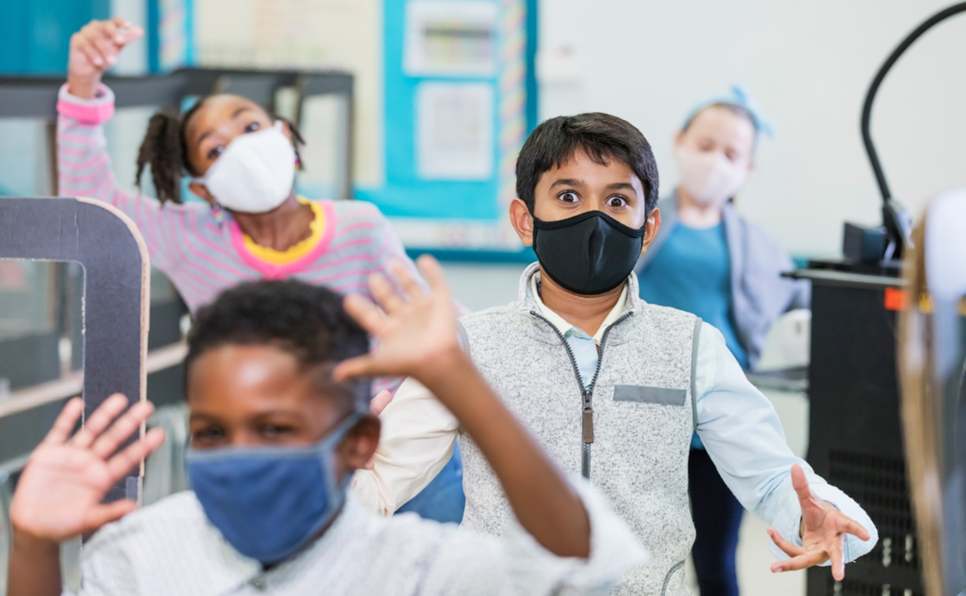A multi-ethnic group of elementary school students in the classroom during the covid-19 pandemic, wearing face masks and standing six feet apart, social distancing. The focus is on the boy wearing a black mask, looking at the camera. He is 9 years old, of Indian ethnicity. The children have playful expressions.
