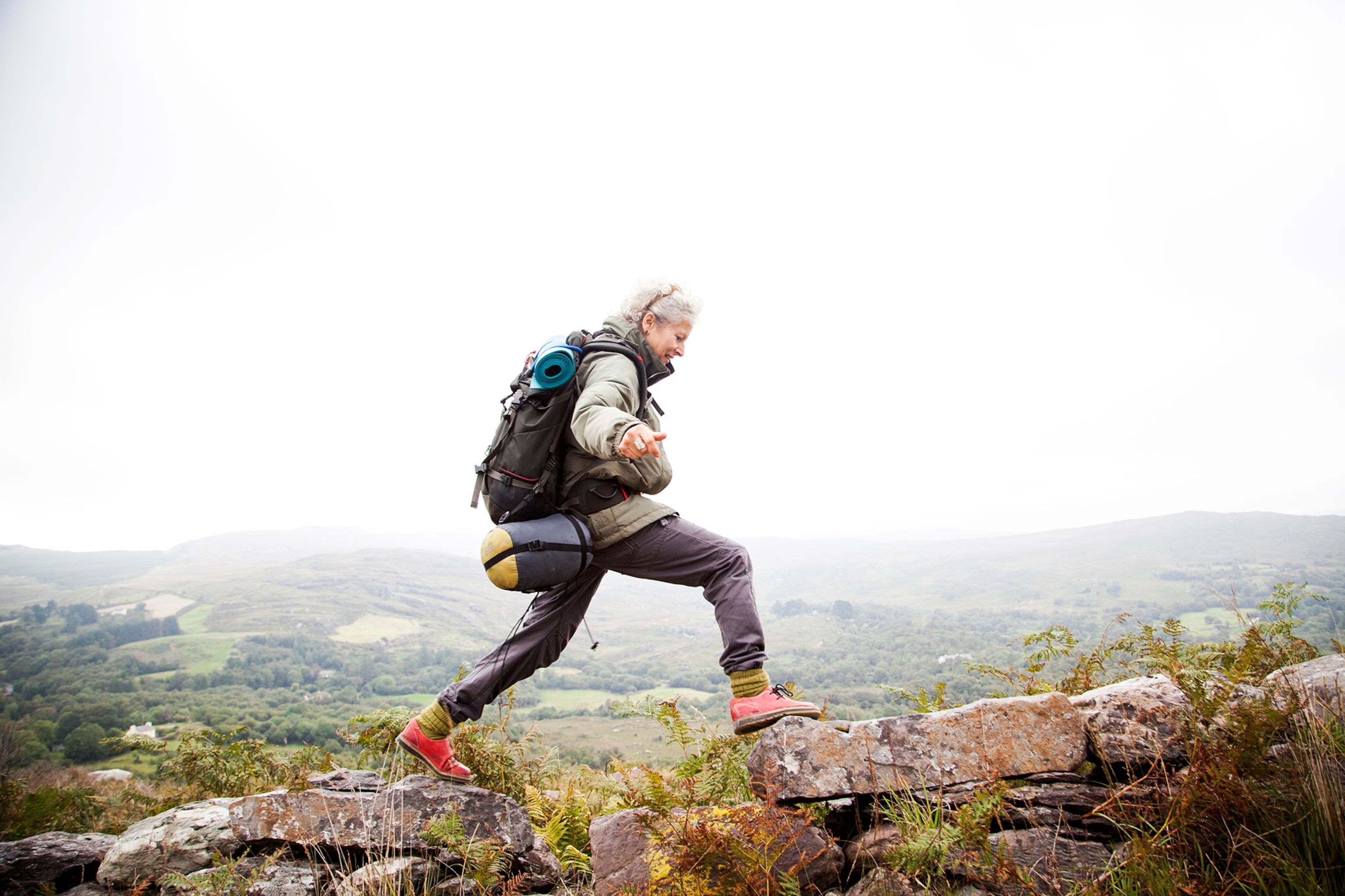 Older woman, with a backpacking back pack, a light grey coat and dark grey pants, happily trekking in the mountains of ireland