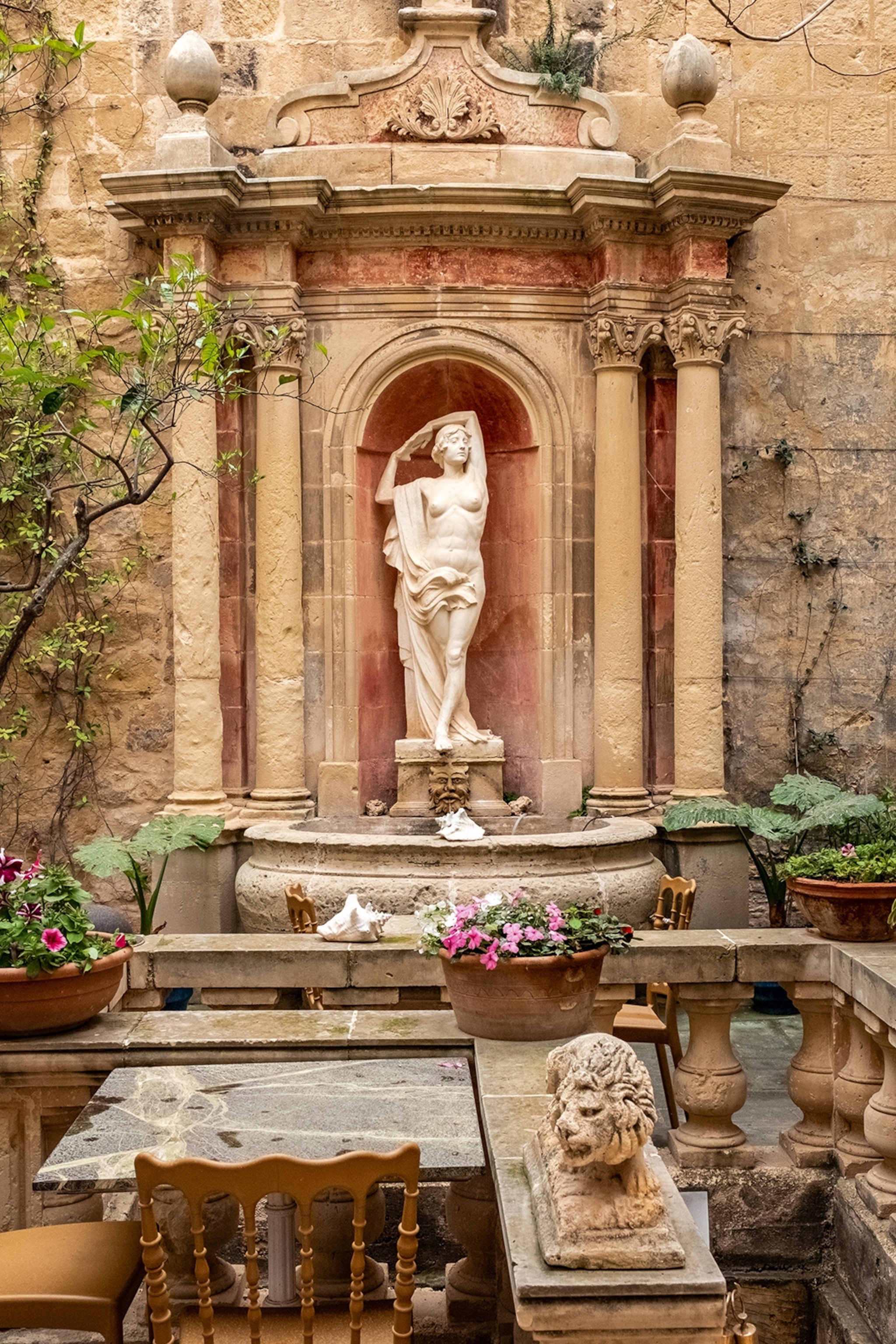 A picture of an old courtyard statue in Valletta, Malta. Planted flower pots are dotted around, whilst the stone architecture is near-immaculate.