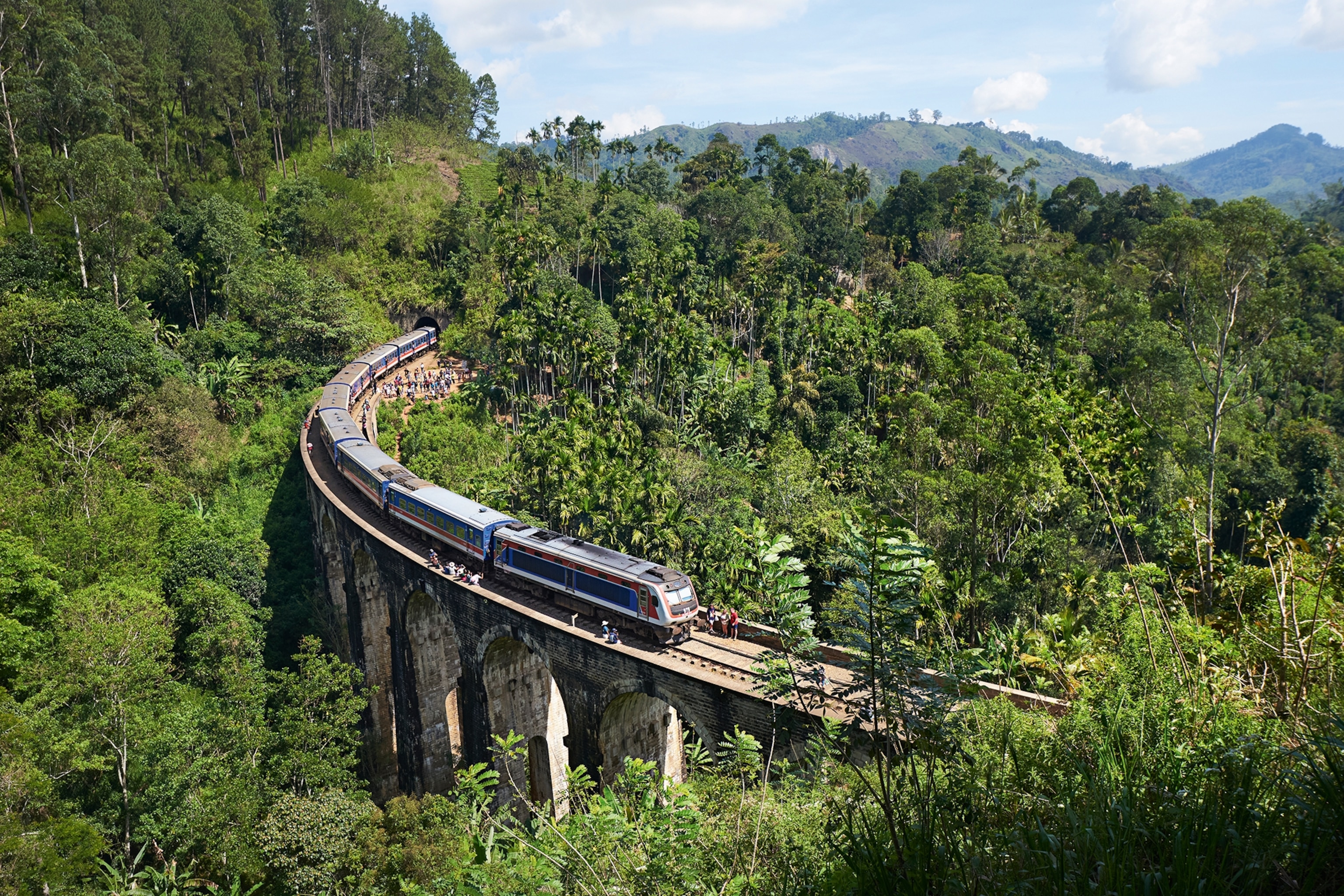 Train passing through a jungle with people sat alongside the tracks
