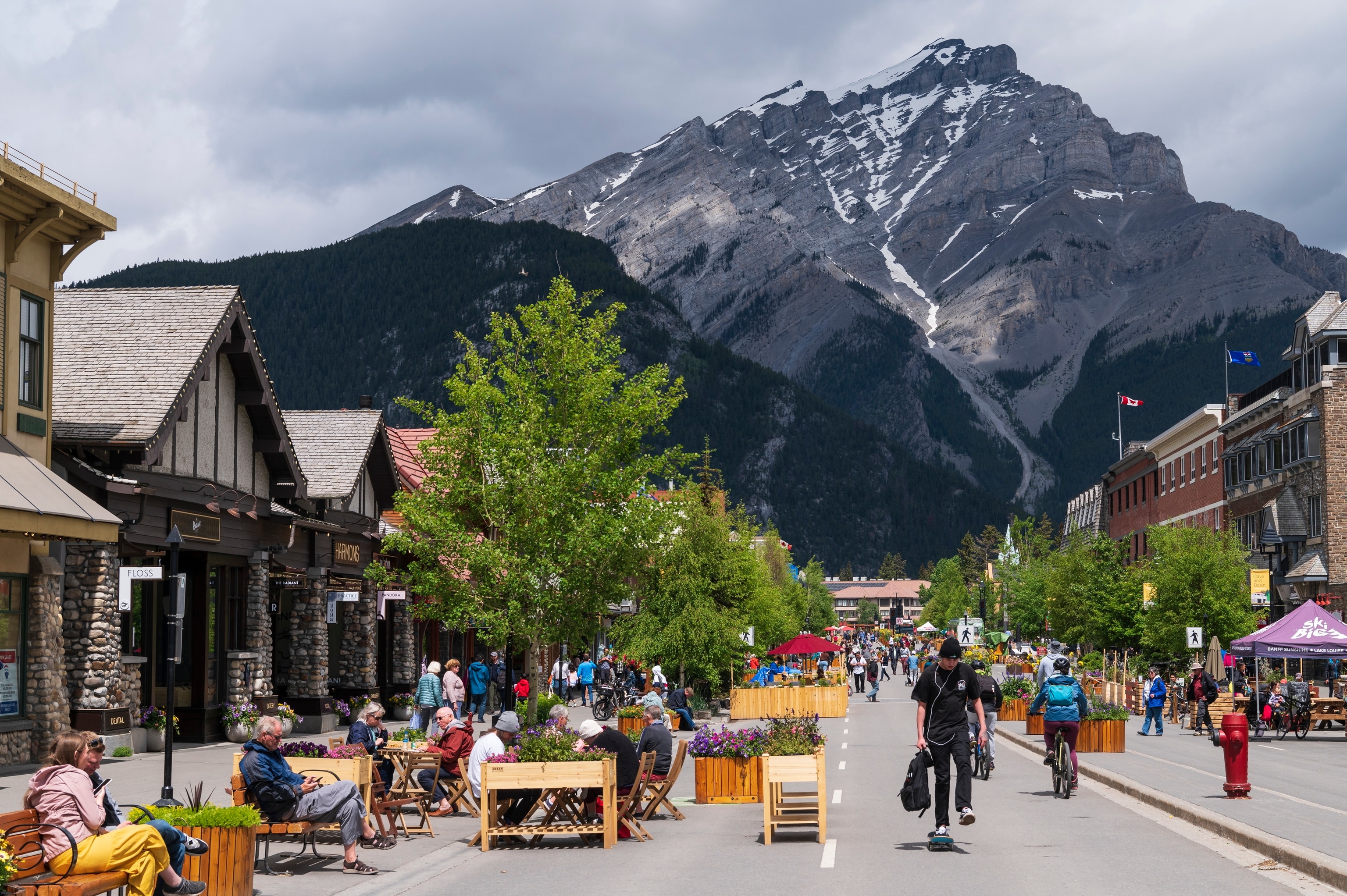 A two-lane road lined with shops cutting through a quaint town is used as a pedestrian zone. In the background, a large, grey, snow-topped mountain looms.