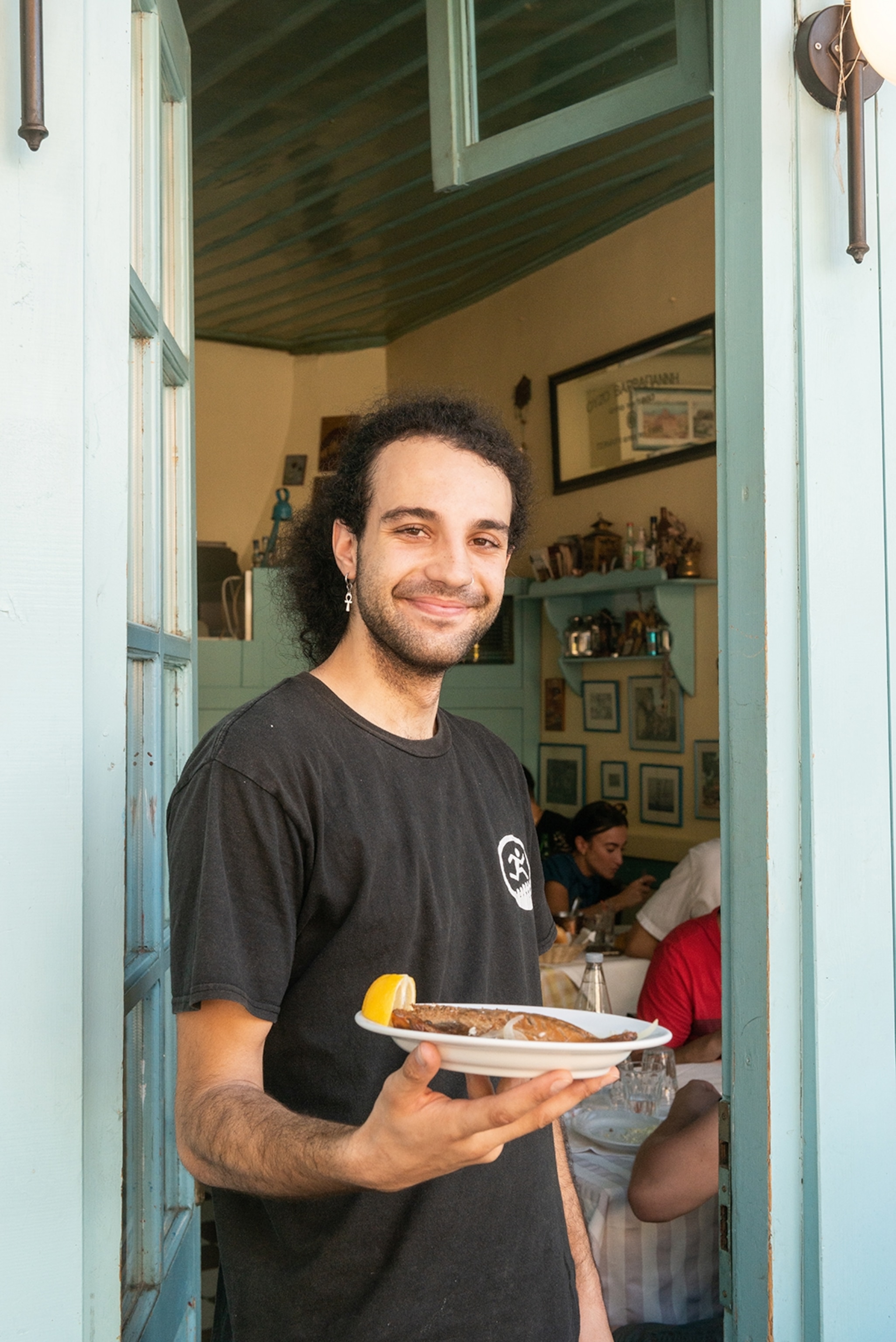 A smiling young Greek waiter in a casual T-Shirt, offering up a plate of fish to the camera, a busy restaurant room behind him.