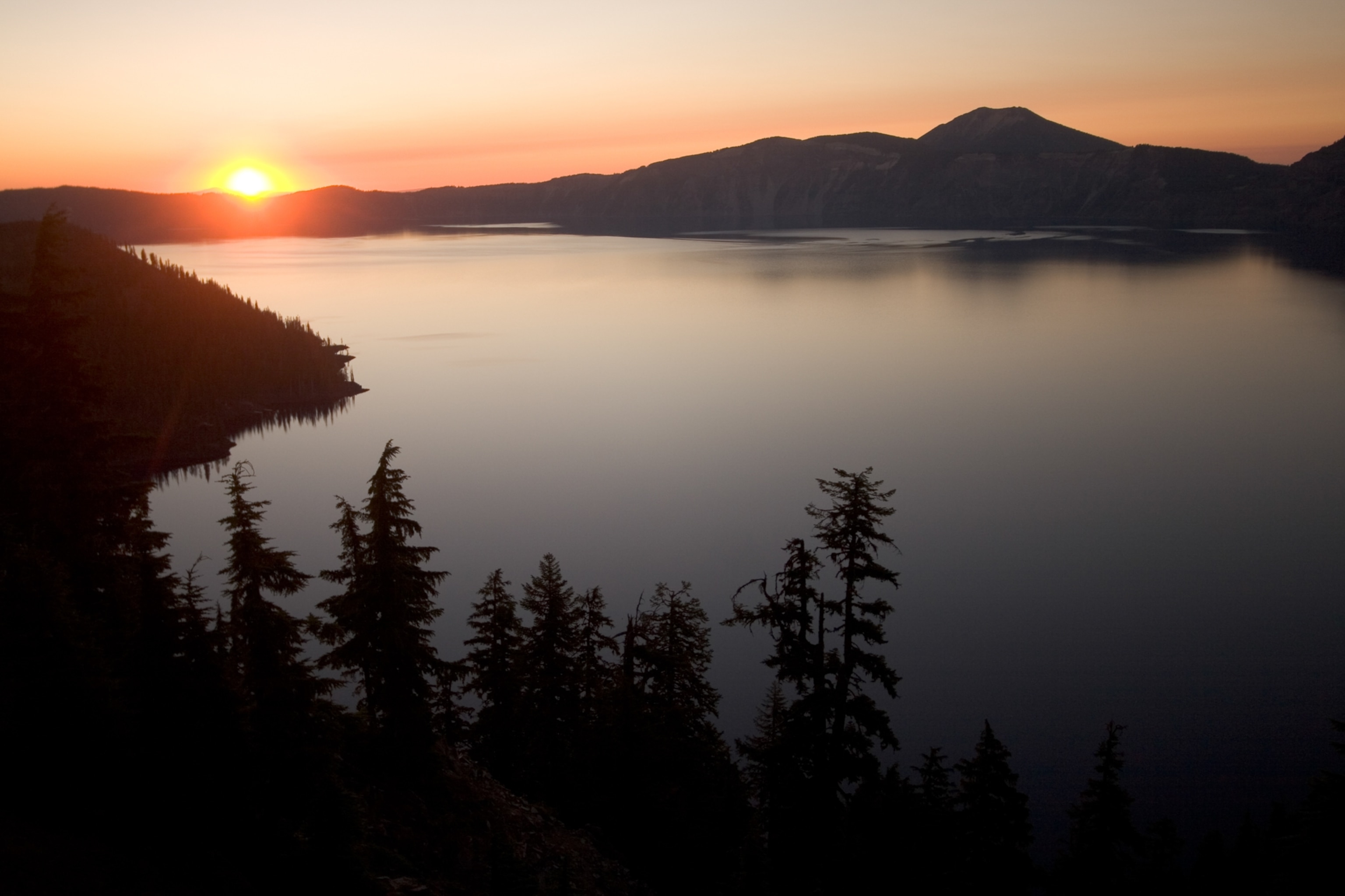 Sunrise over Crater Lake, with the foreground trees and mountains in the background in silhouette against the orange sun.