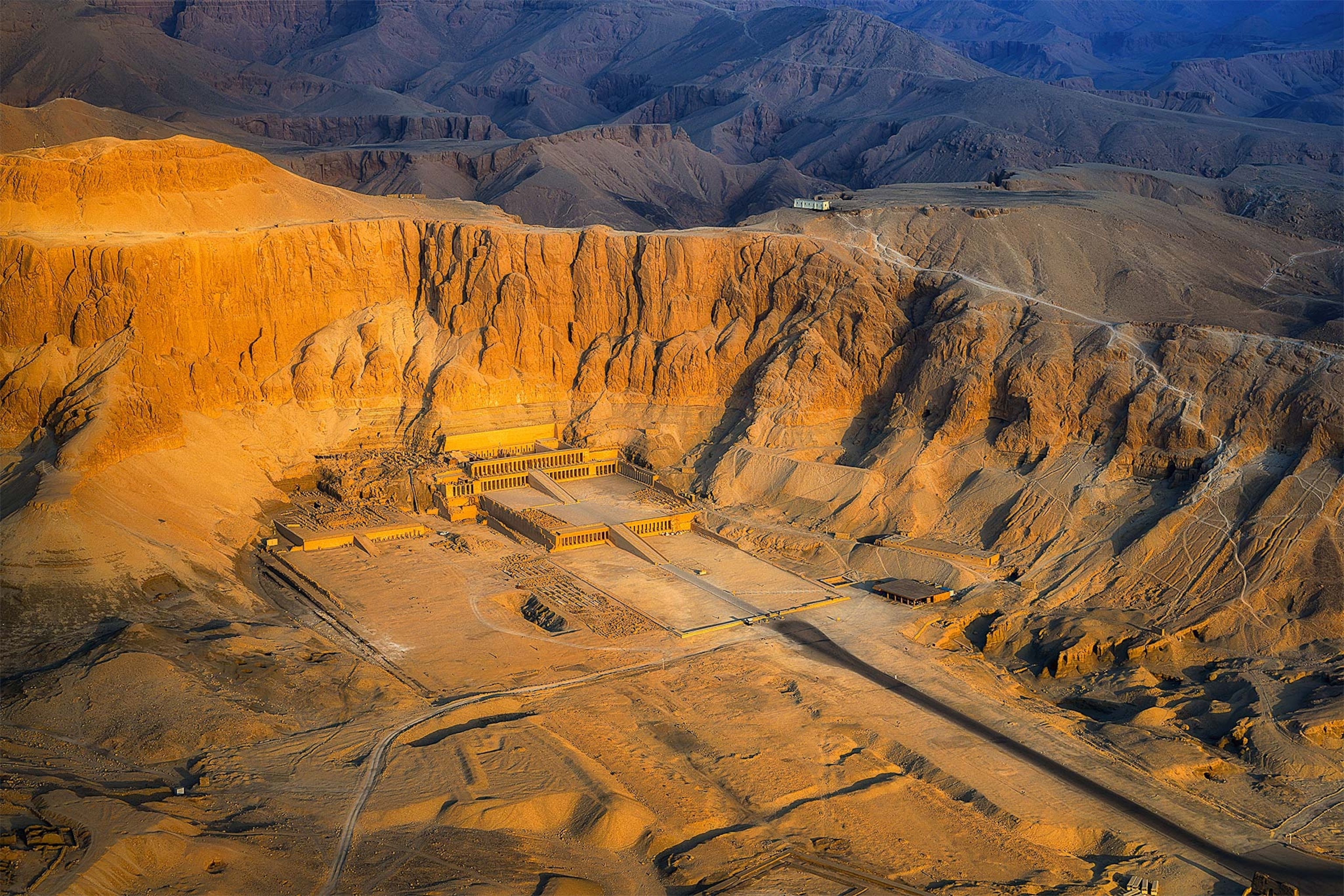 An aerial view of the Deir el Bahri temple site, in the desert, with a sun setting casting orange light on the sand