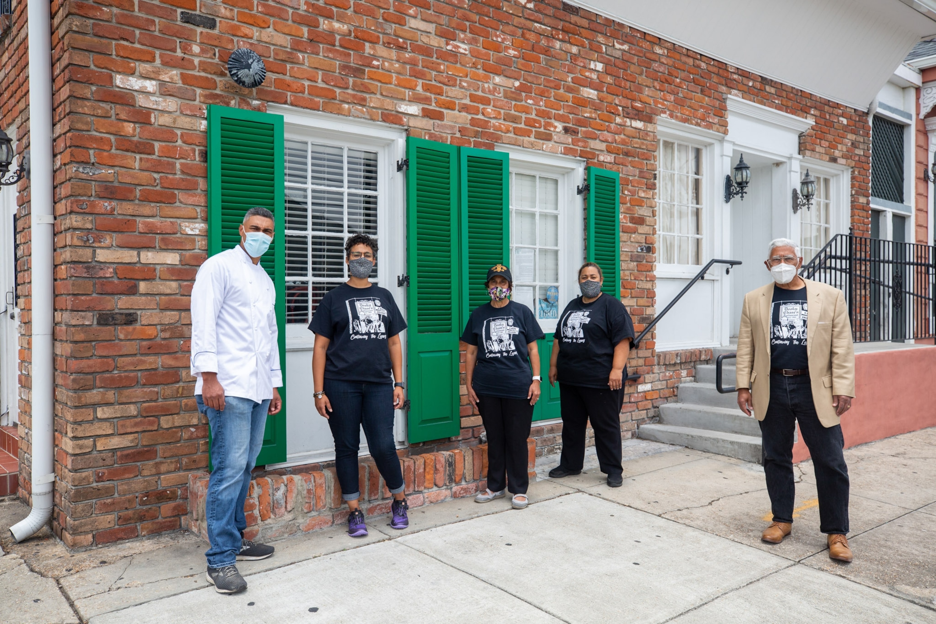 five adults with masks on standing outside of a brick building