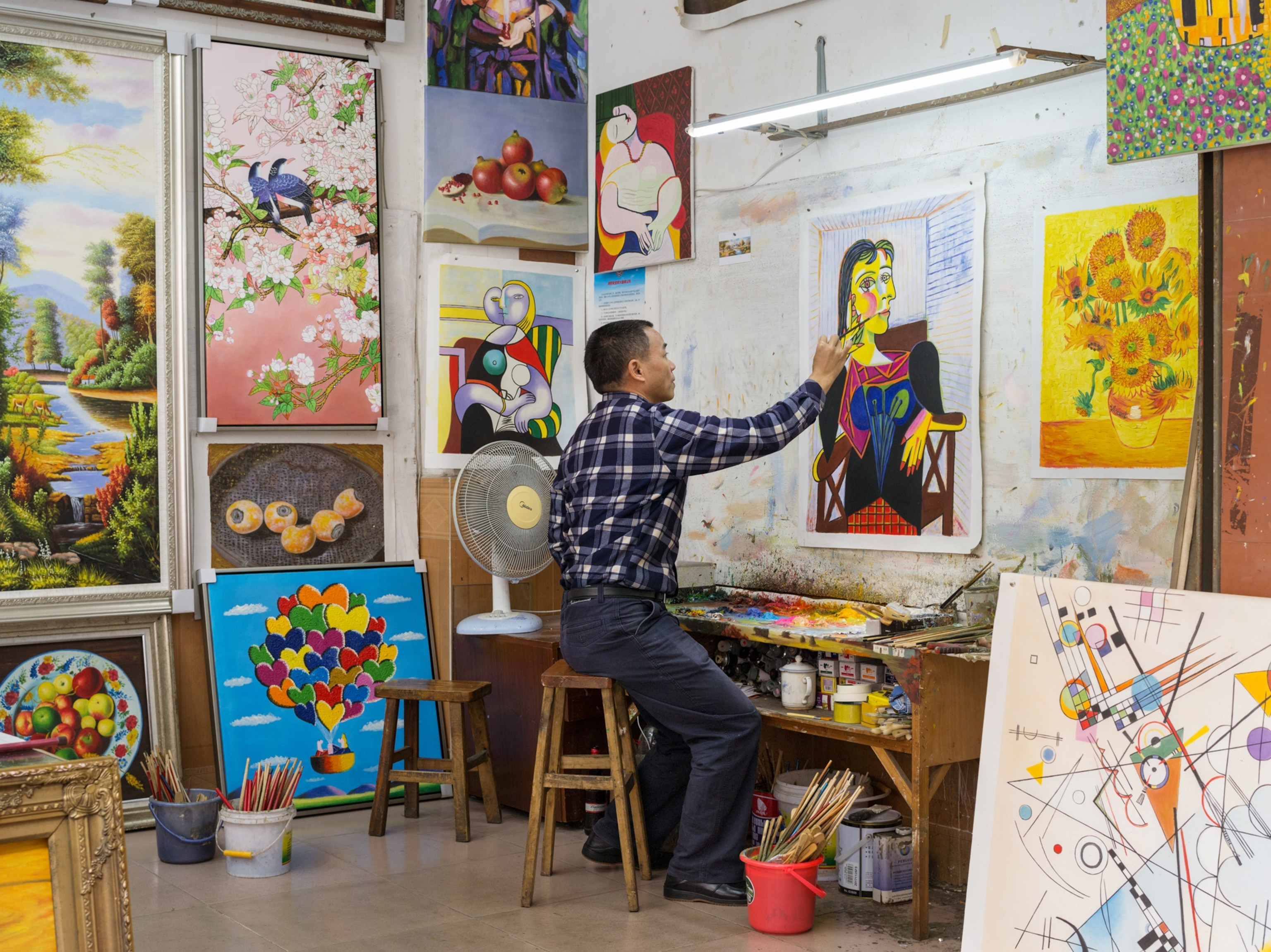 a man sitting on a stool painting a Picasso surrounded by other paintings